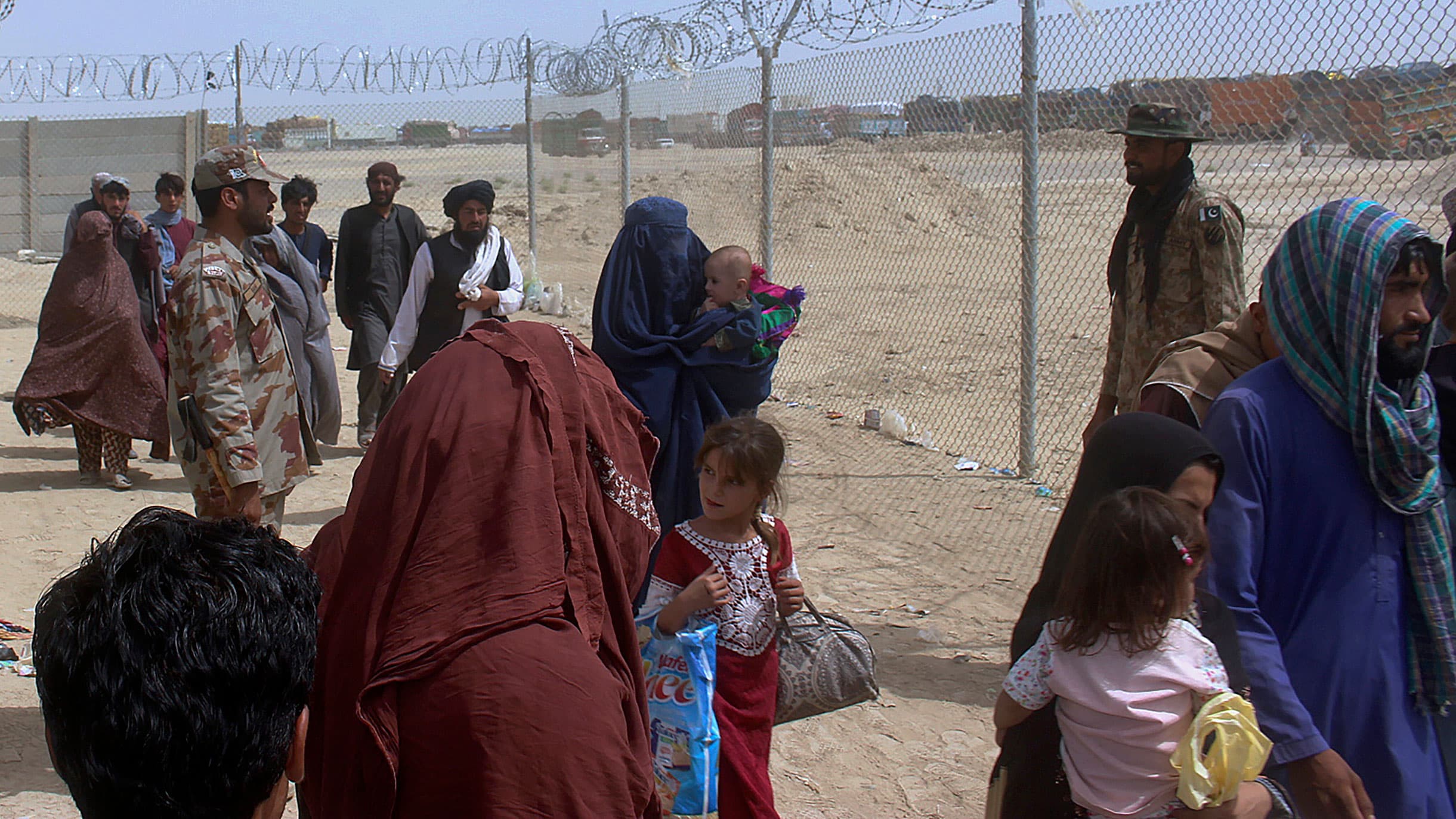 Soldiers are shown standing guard near a barbed-wire fence with a larger crowd of people shown standing and waiting.