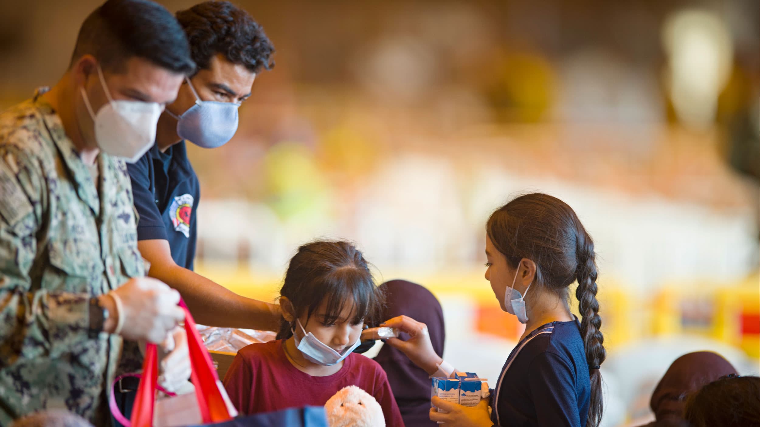 Two men are shown wearing medical face masks and handing out drink boxes and snacks to two young Afghan refugees.