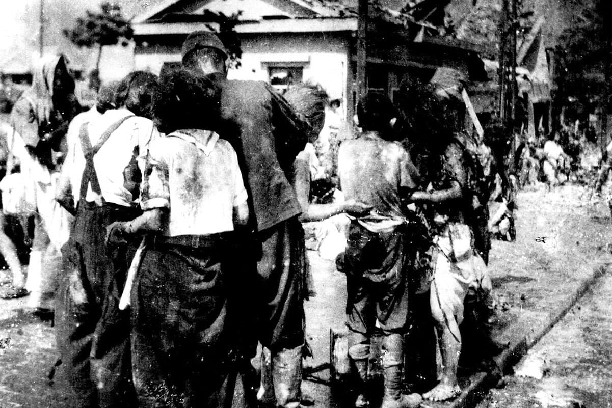 A crowd of survivors are shown standing and sitting on a bridge with several damaged buildings in the distance.