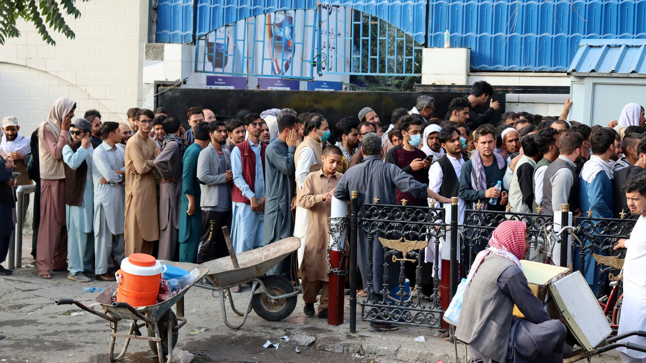 A large crowd of people are shown standing, packed in a line with a blue-sided building in the background.