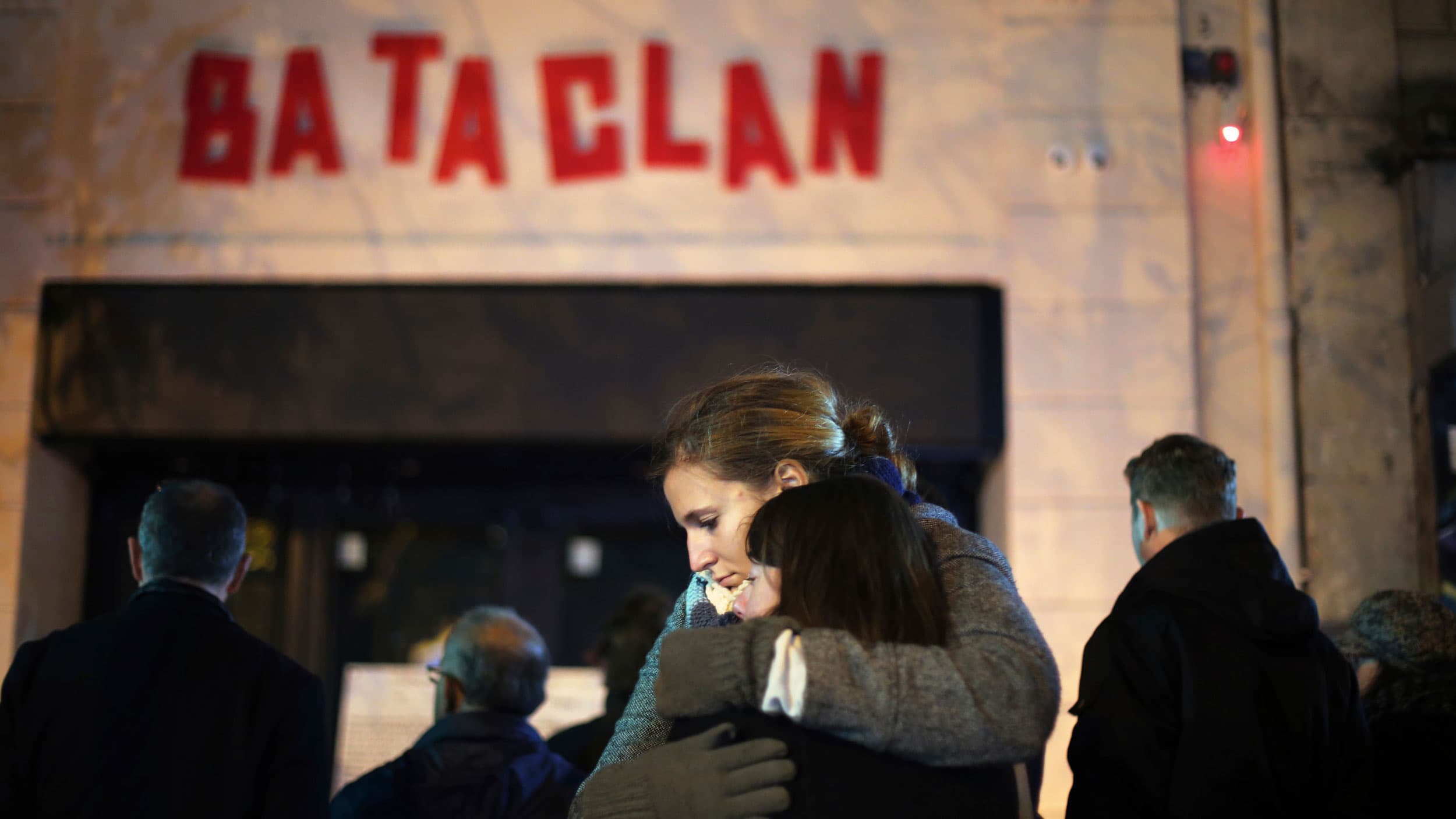 Two women are shown hugging each other with the entrance and red-lettered sign of the Bataclan concert hall in the background.