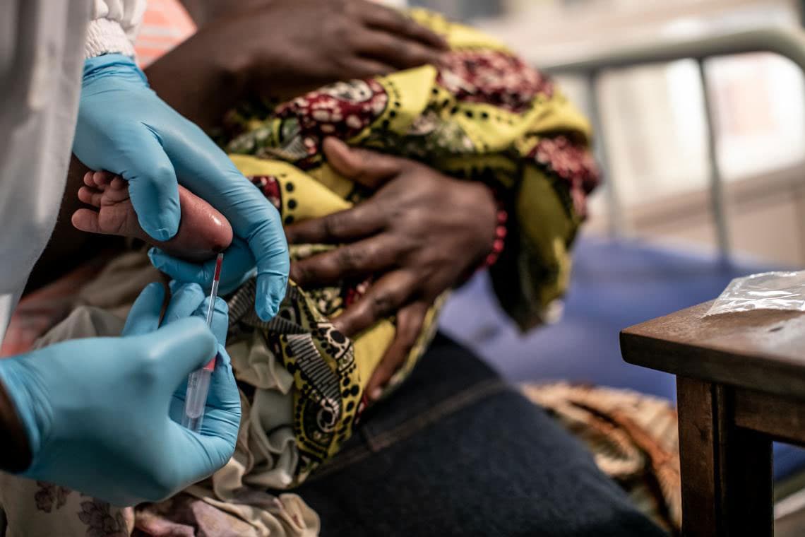 A baby's foot is pricked for a blood test by a pair of hands wearing blue gloves.