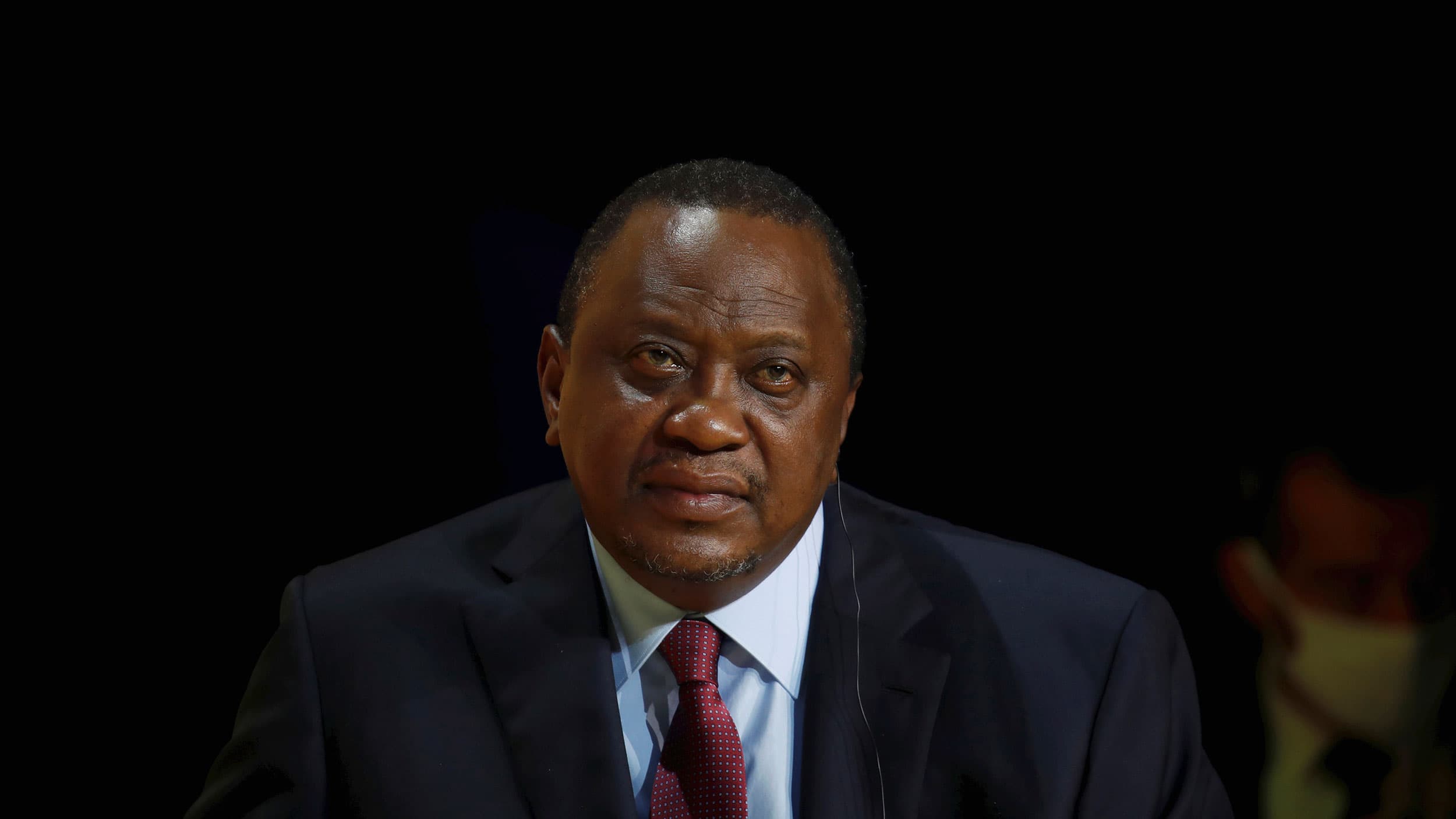 Kenya's President Uhuru Kenyatta is shown sitting and listening while wearing a dark suit and red tie.
