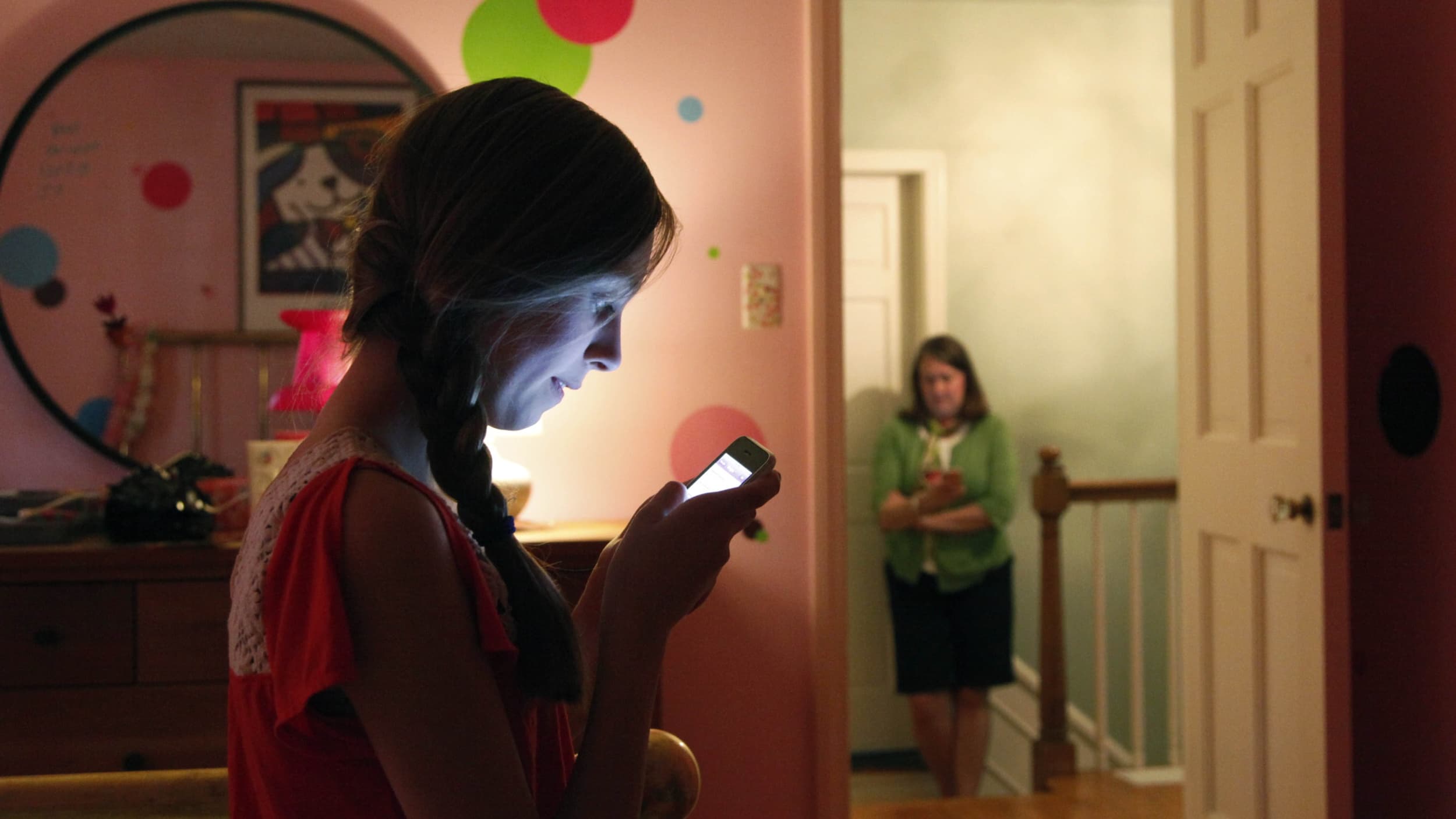 A woman sits on a bed using her phone with another woman in the hallway doing the same