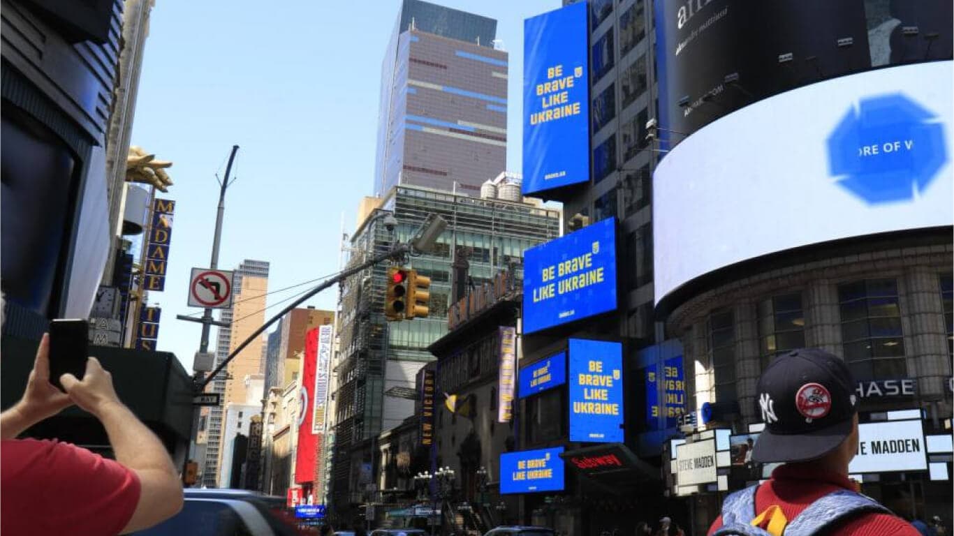 People's hands are shown holding phones and cameras, pointed at three blue billboards that say in yellow 'be brave like Ukraine'