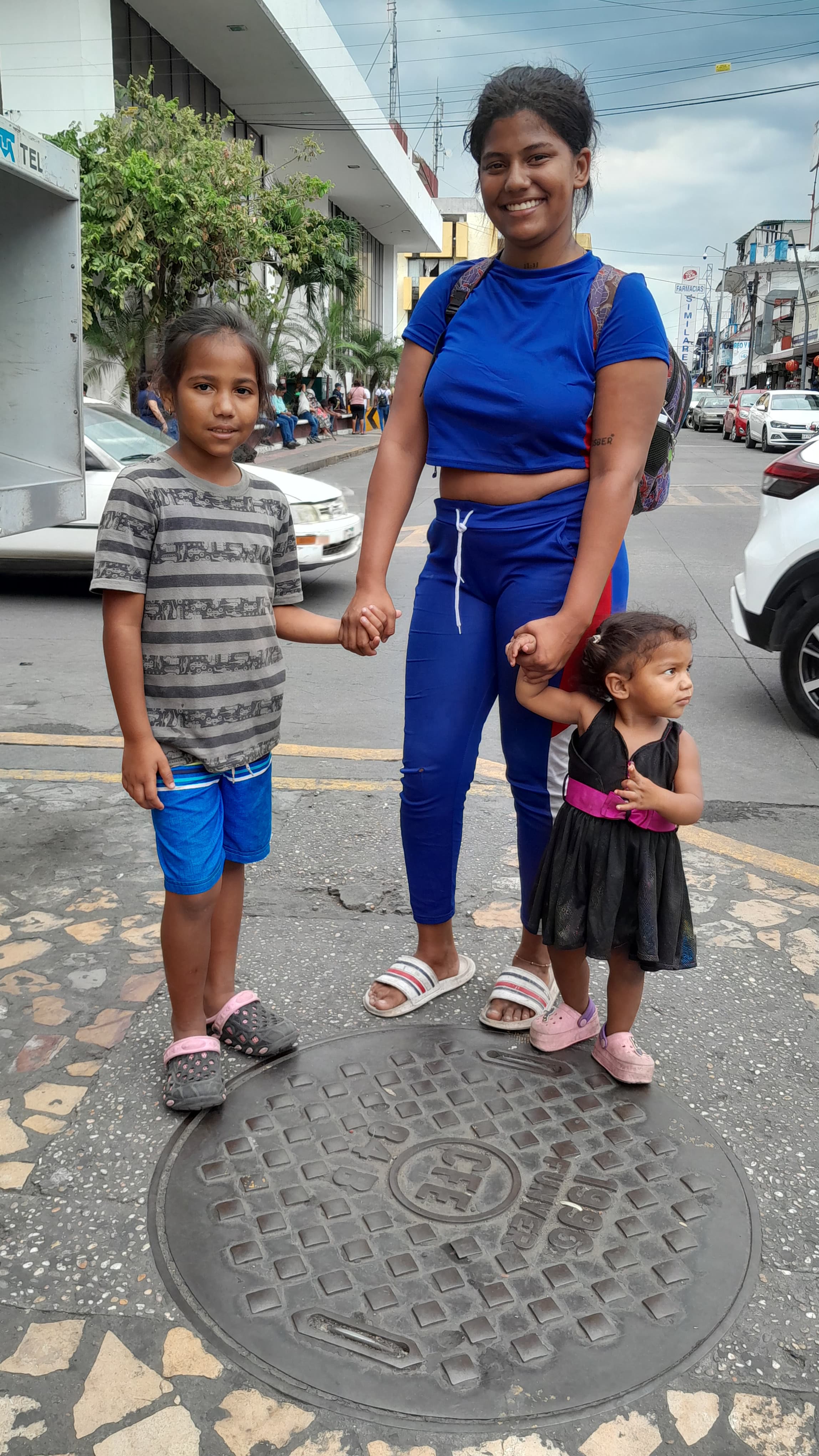 A woman wearing blue a top and pants, holding holding the hands of her two kids, a small girl and tall boy, on each side of her.