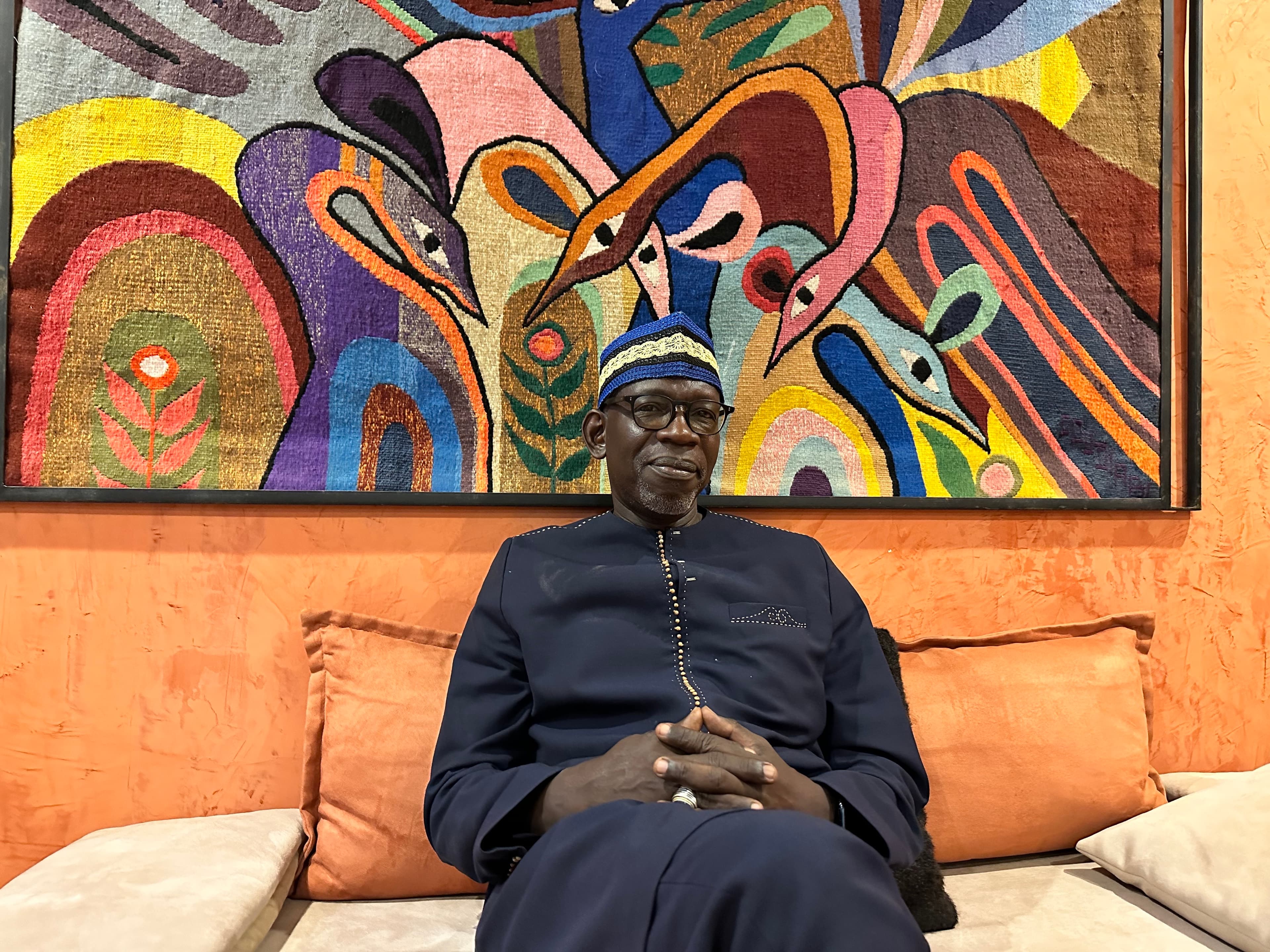 a man wearing traditional African clothing, sitting down in front of colorful artwork