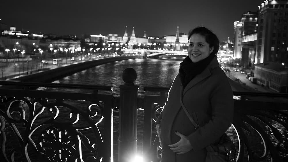 a black and white photo of apregnant woman holding her belly and wearing a large coat standing in front of a bridge