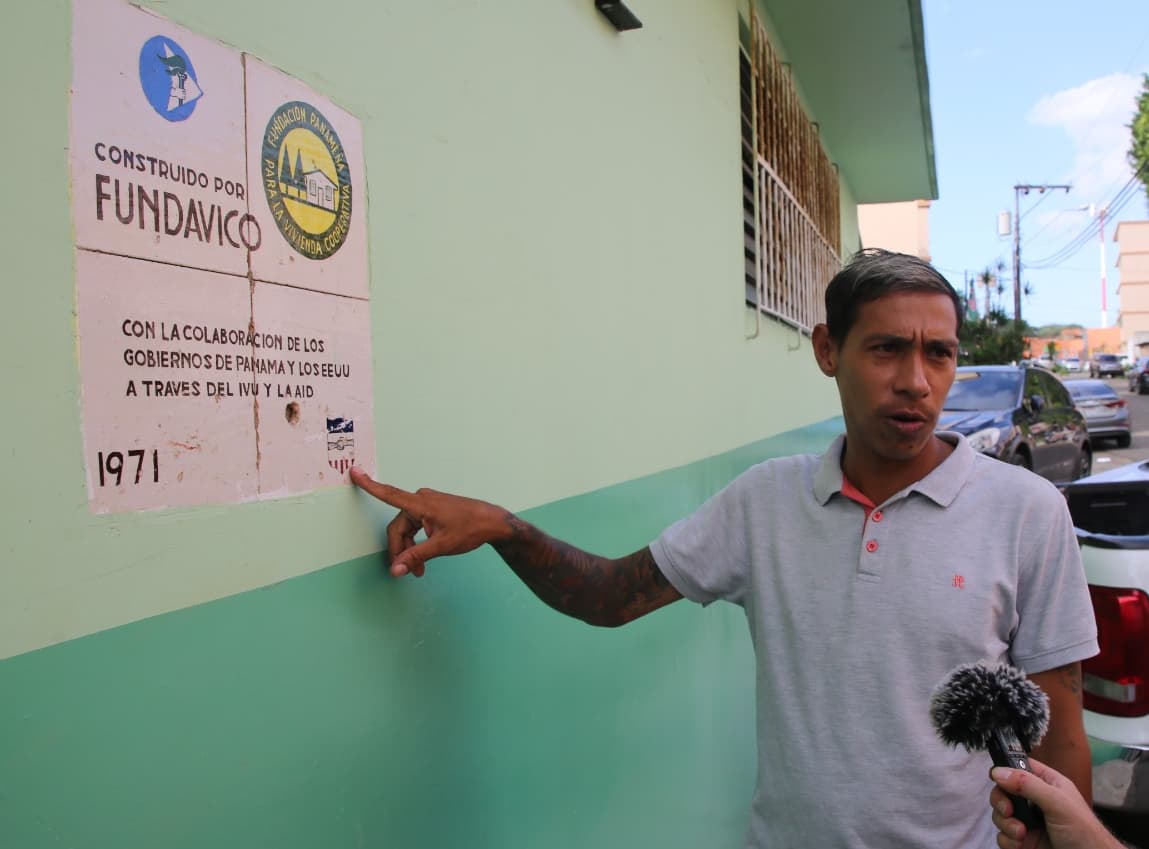 A man in a white collared shirt points to a bullet hole in a wall