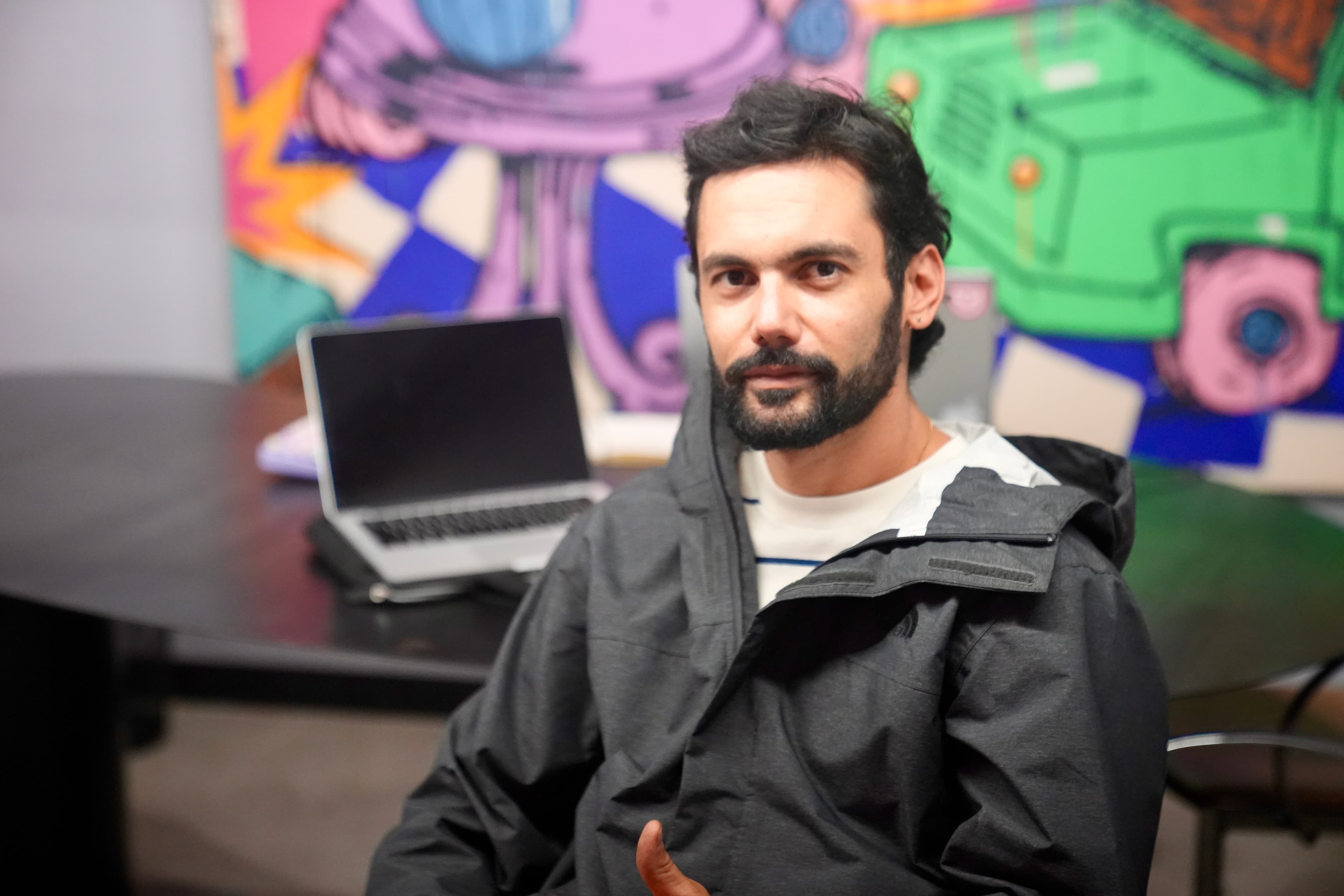Potrait of a man sitting down in a black jacket in front of a colorful mural in the background