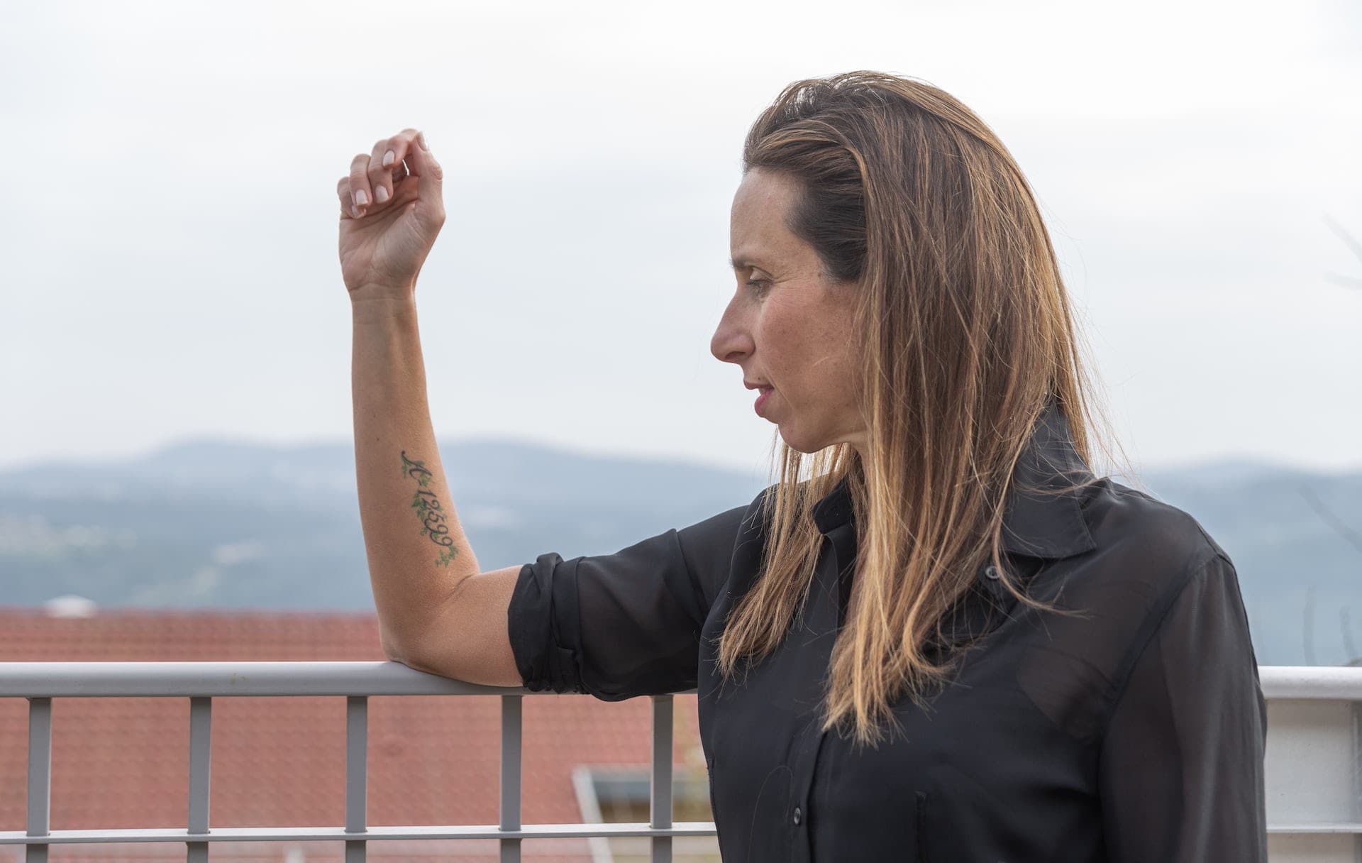 A woman in a black shirt holds up her arm on which there is a tattooed number.
