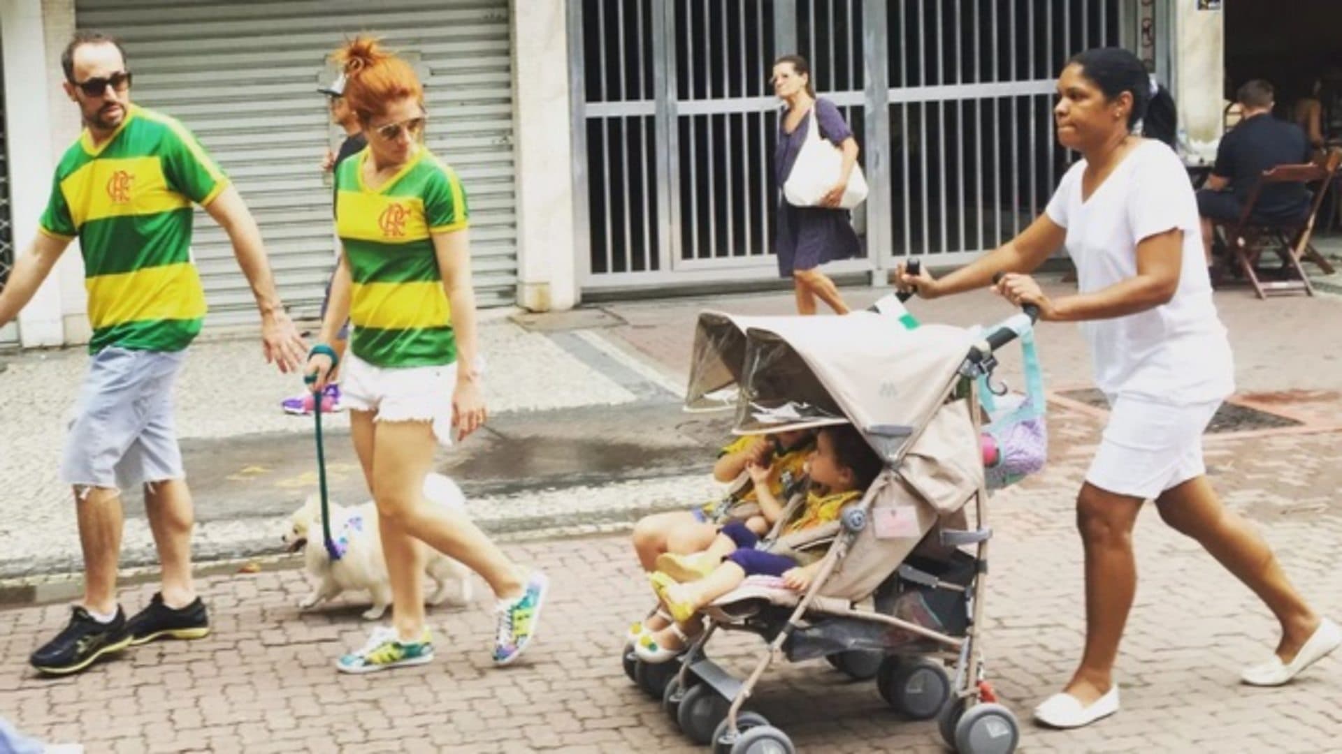 A white couple look back at their two children in strollers, being pushed by their black nanny
