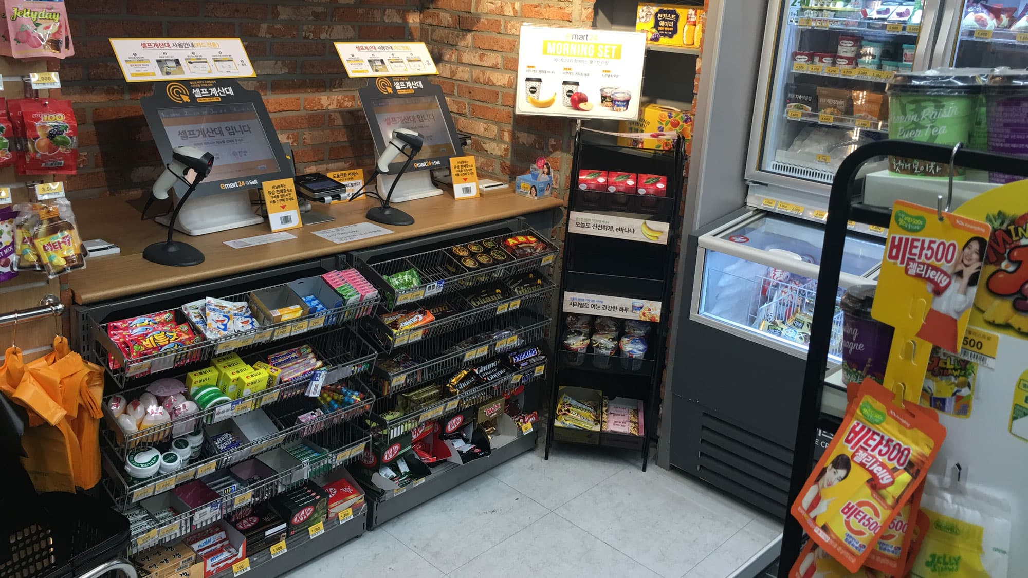 Two computers with scanners are set up on a counter in a convenience store, surrounded by packaged snacks and rows of gum and mints.