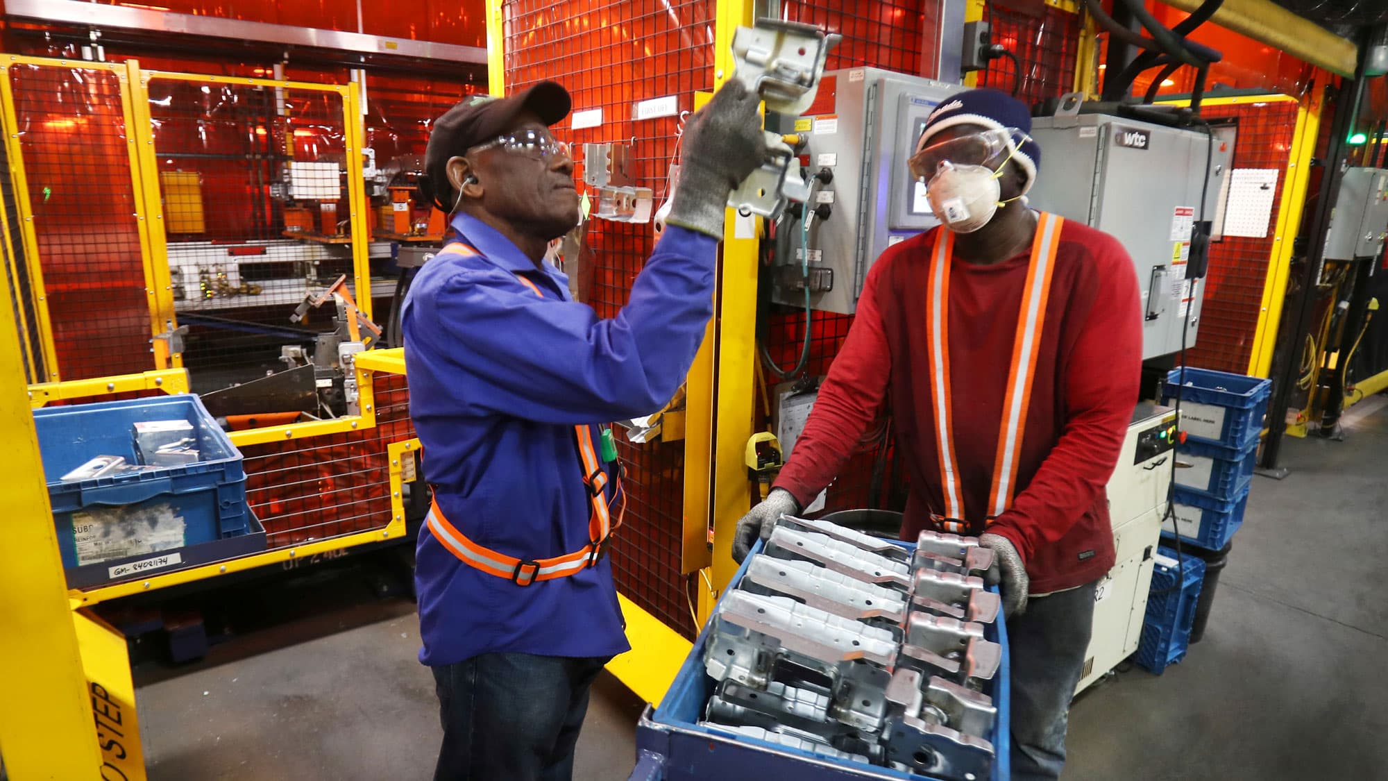 One person inspects a piece of metal, while another person holds a cart containing a more of the same pieces of metal.