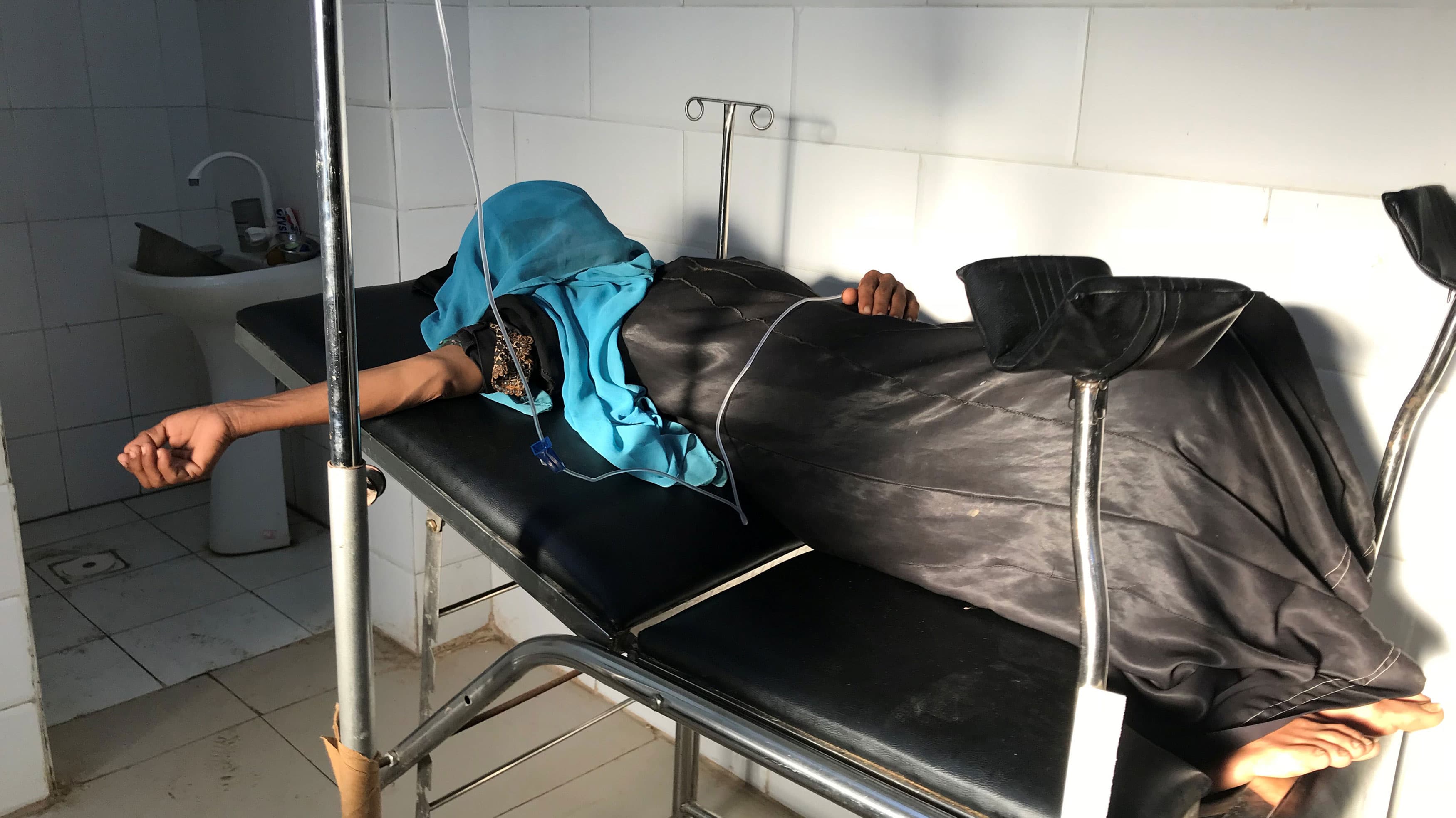 A woman lies on a table, her arm outstretched. Her face is hidden by a blue cloth.