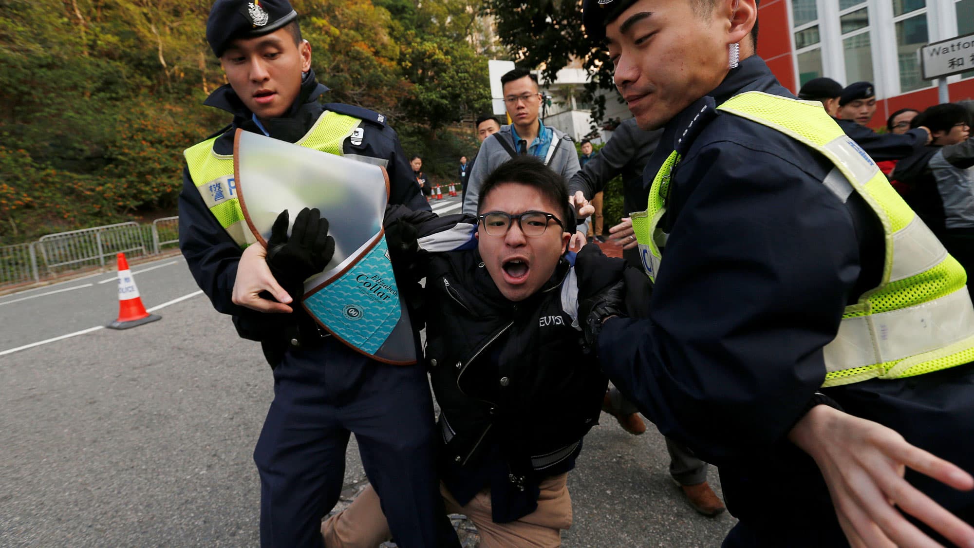 Two police in yellow vests hold the arms of another man, who is yelling as he is being dragged by police.