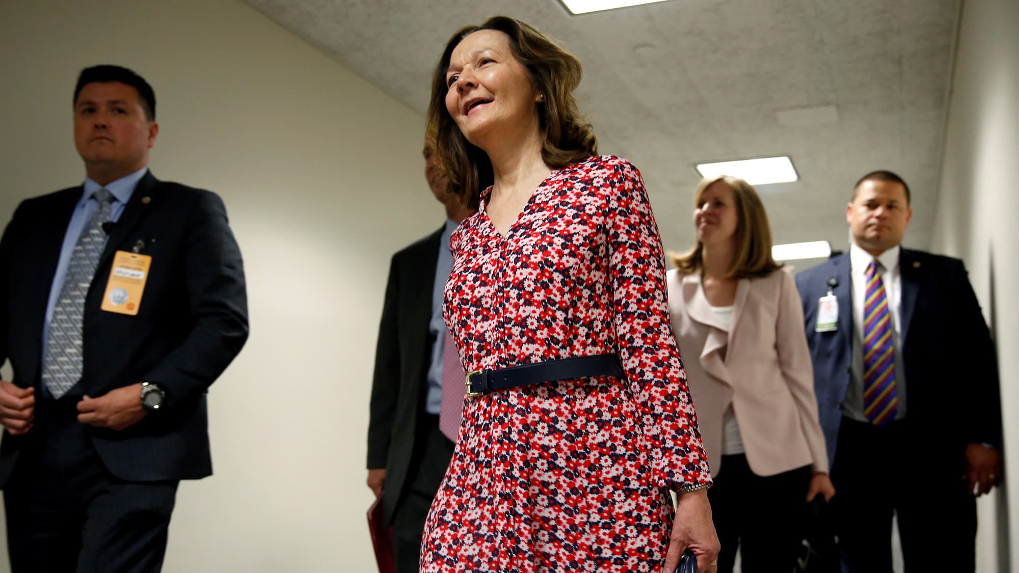 A woman in a red dress walks down a hallway flanked by men in suits.