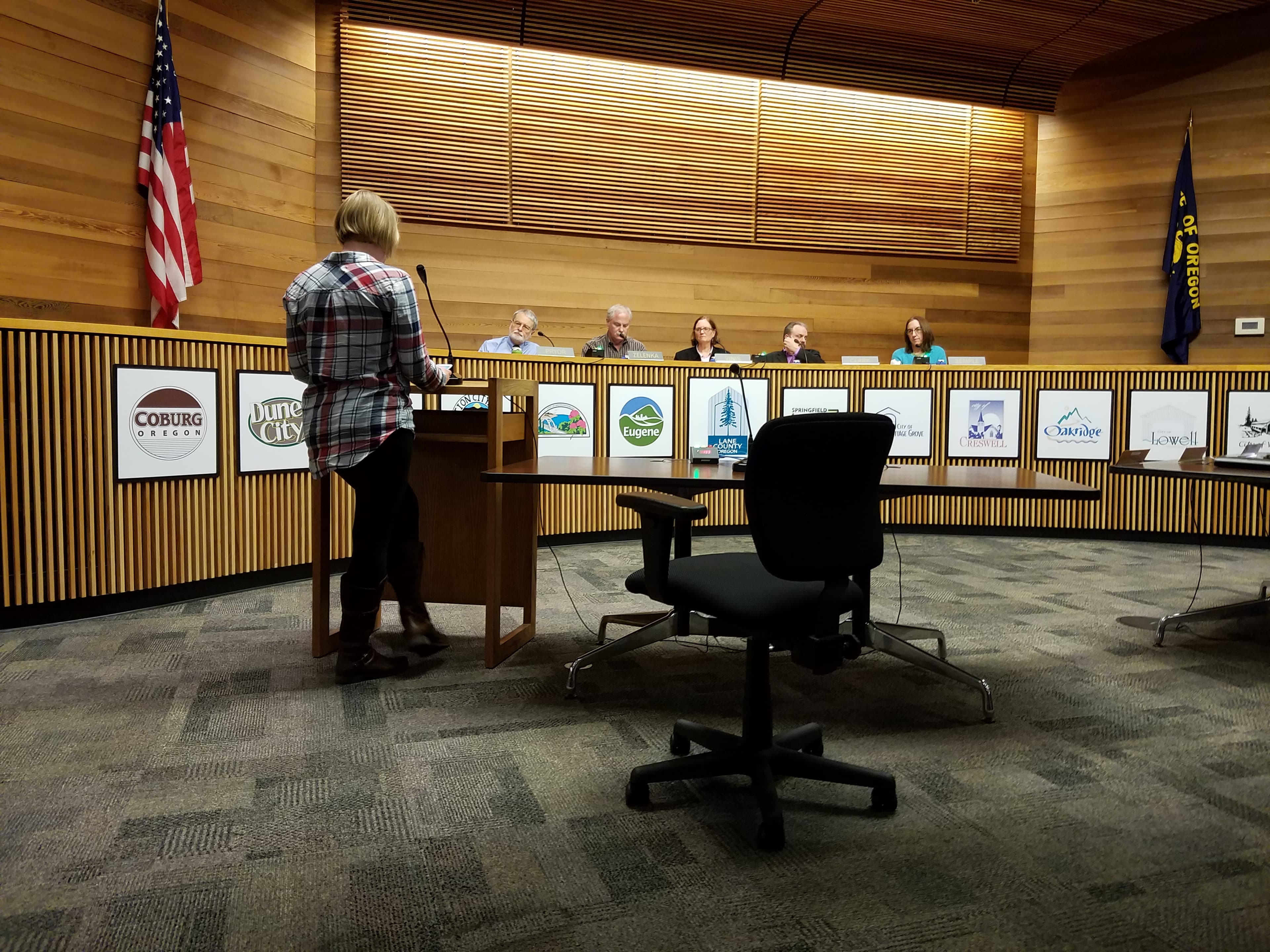 Woman, photographed from behind, stands at a podium in front of panel of people