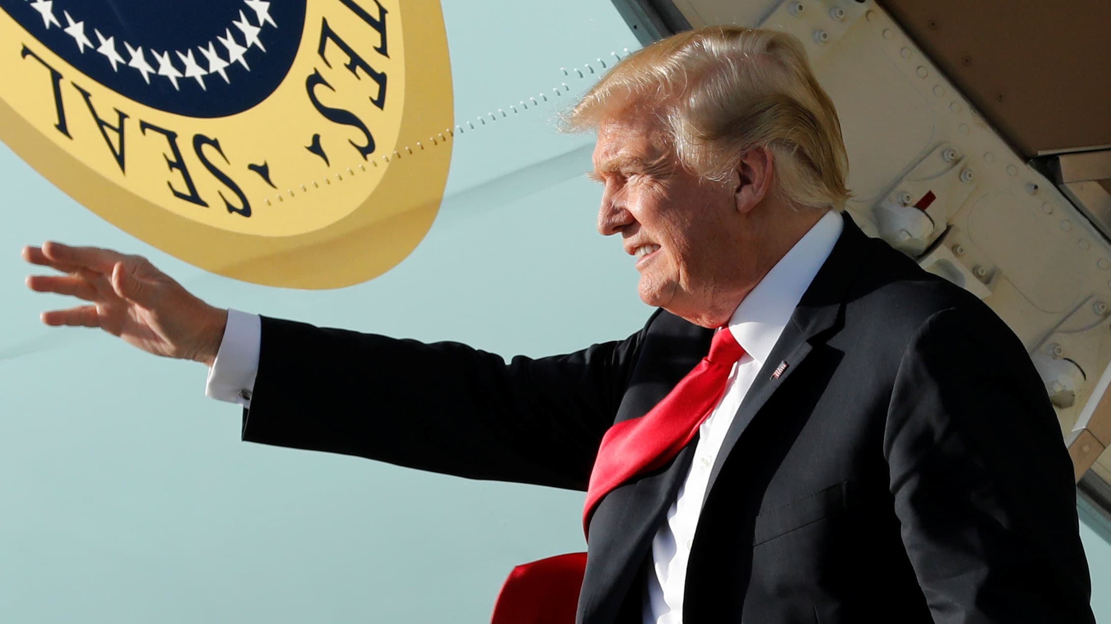 Donald Trump stands on the steps of Air Force One waving with the seal of the president to his right.