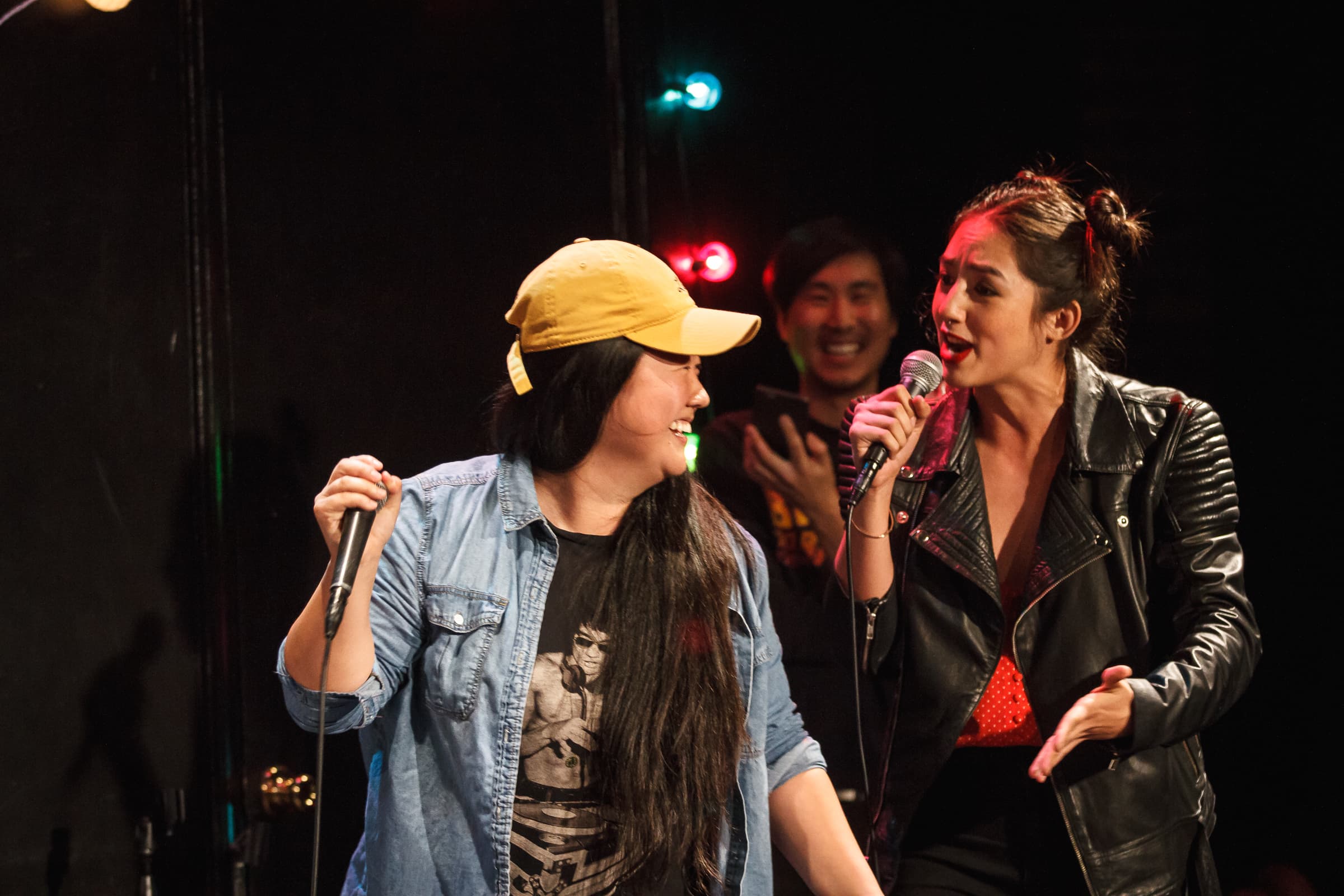 Two female performers are standing in front of each other holding microphones.