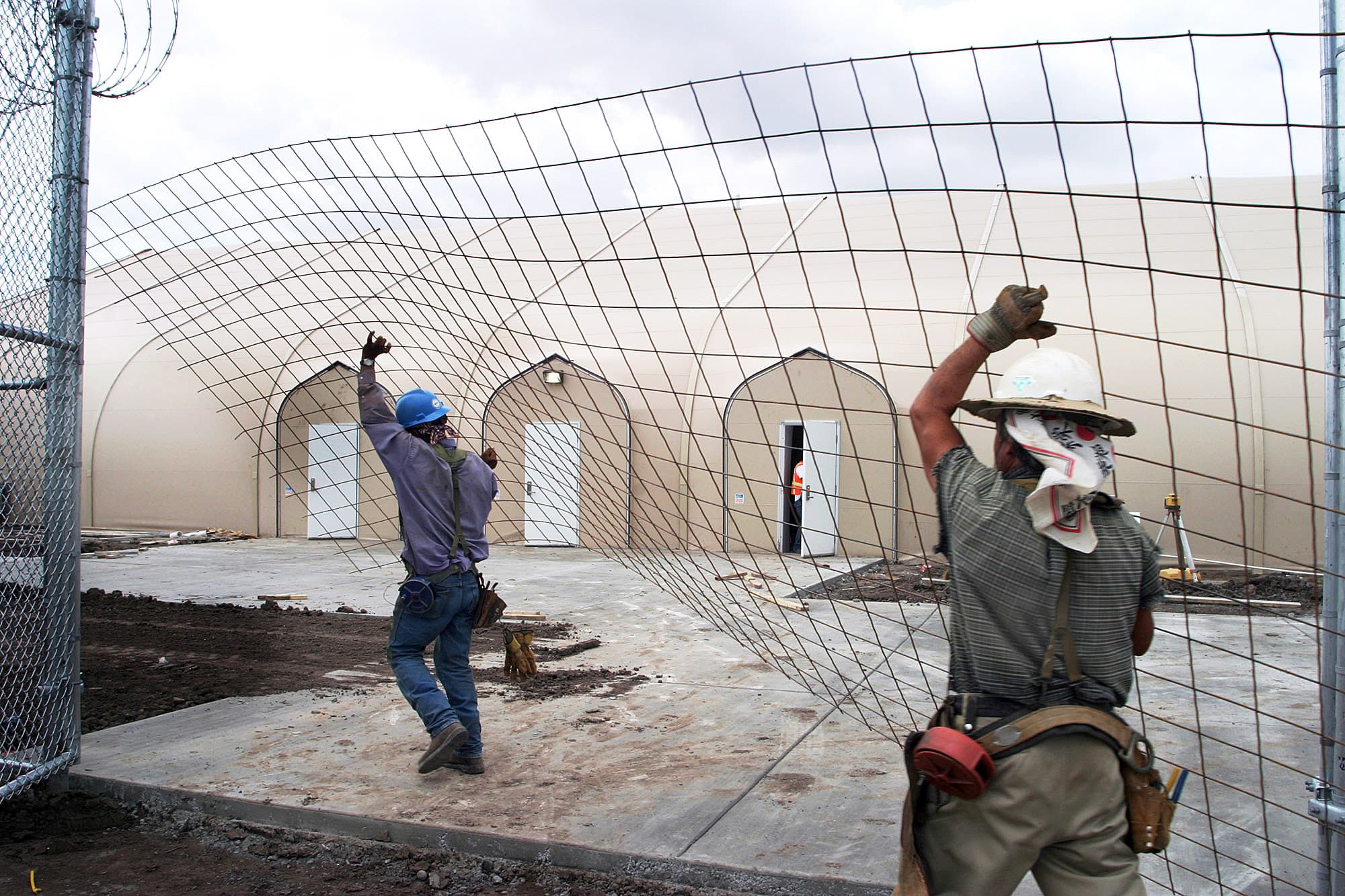 Workers raise a fence in front of white kevlar tents