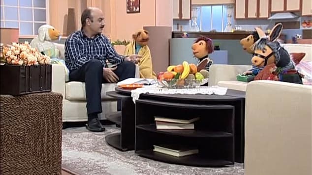 Mr. Host reading a book with the puppets of Kolah Germezi, including the sheep Babayi.