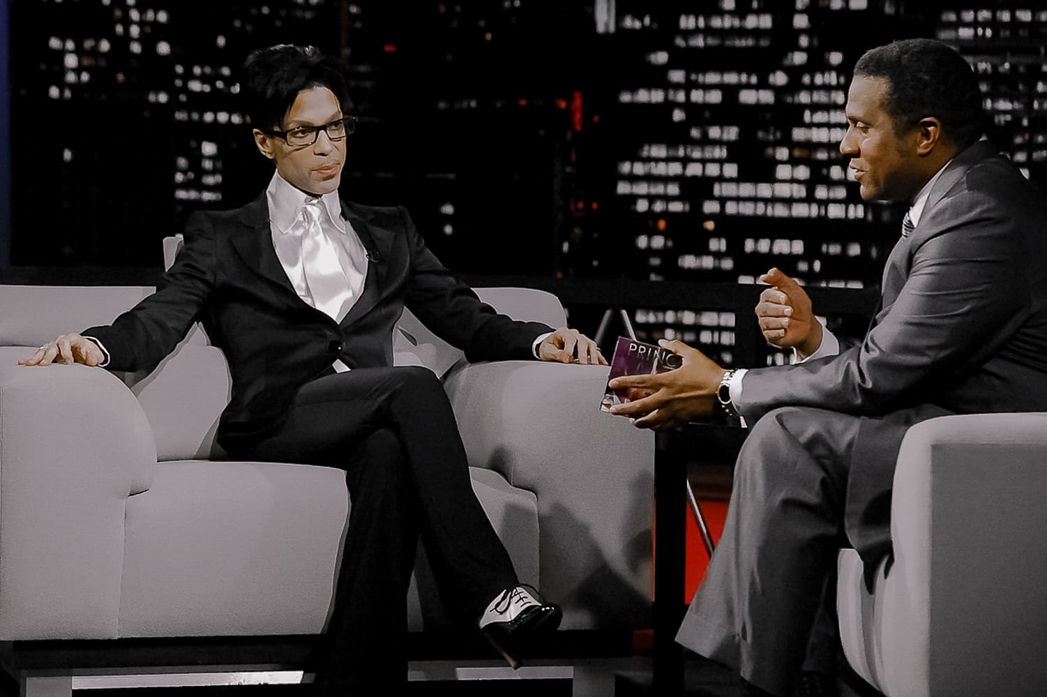 Prince, cross-legged on the set of the Tavis Smiley Show, looks at Smiley as he asks a question