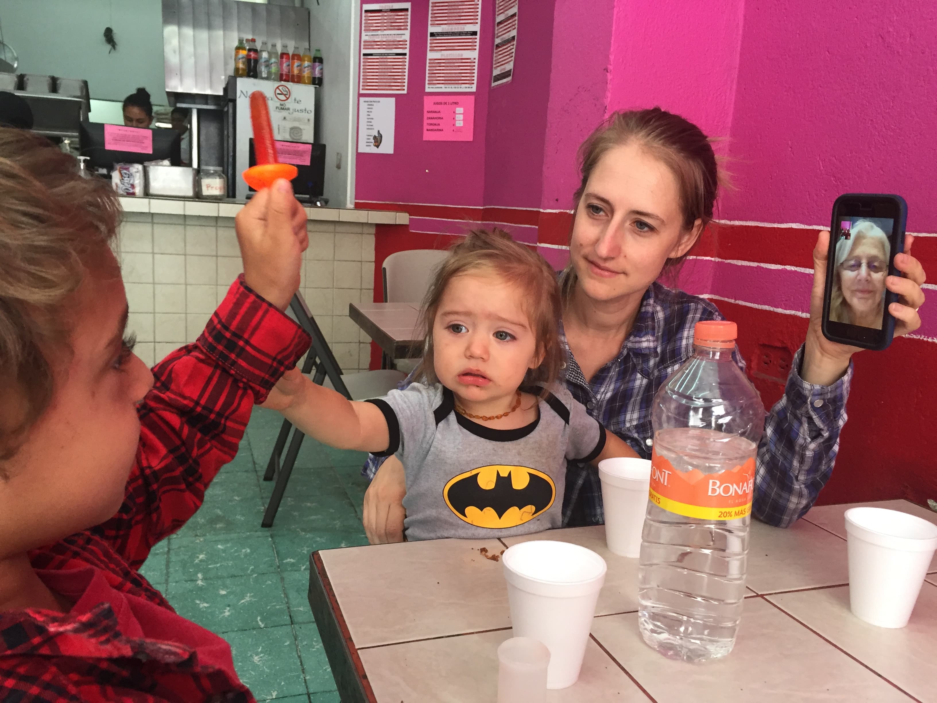 Woman sitting at table with two small children, holding up phone with another woman on video call