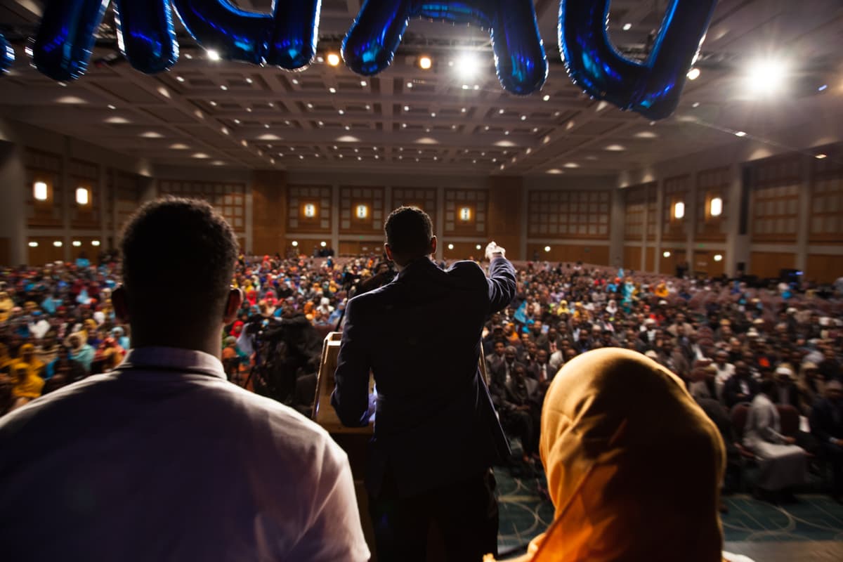 Man standing on stage looking out at crowd, photographed from behind