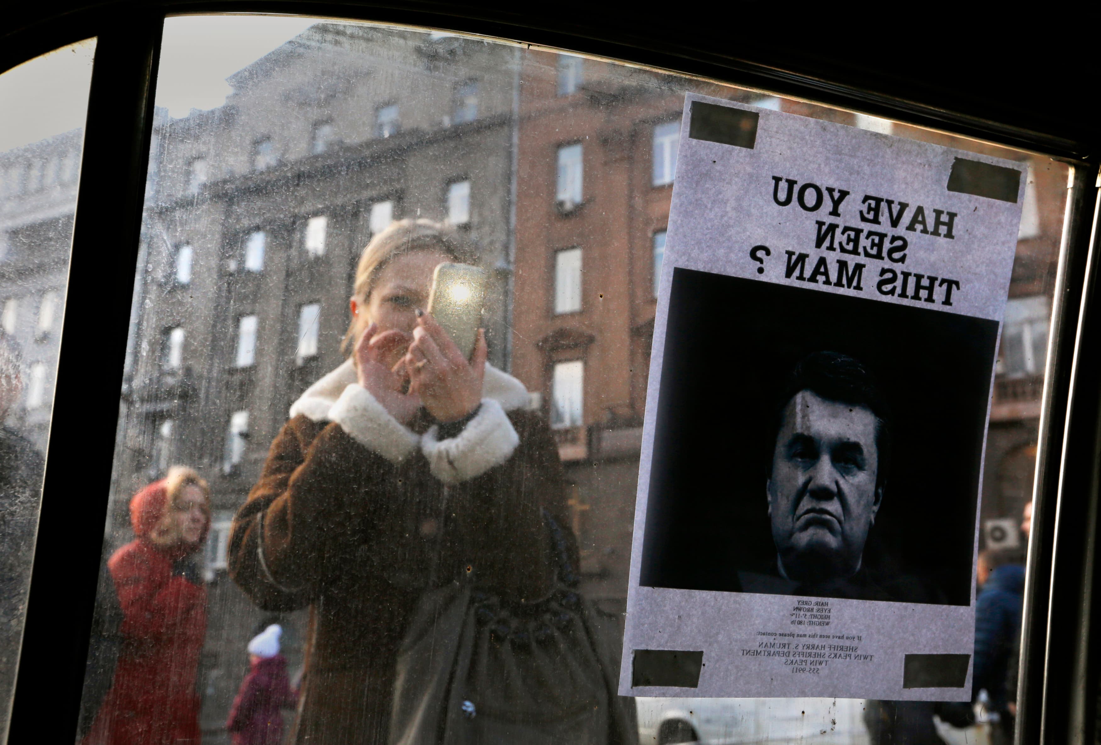 A woman takes photos of a "Wanted" notice for fugitive Ukrainian President Victor Yanukovich, plastered on the window of a car used as a barricade, near Kiev's Independent Square February 24, 2014.