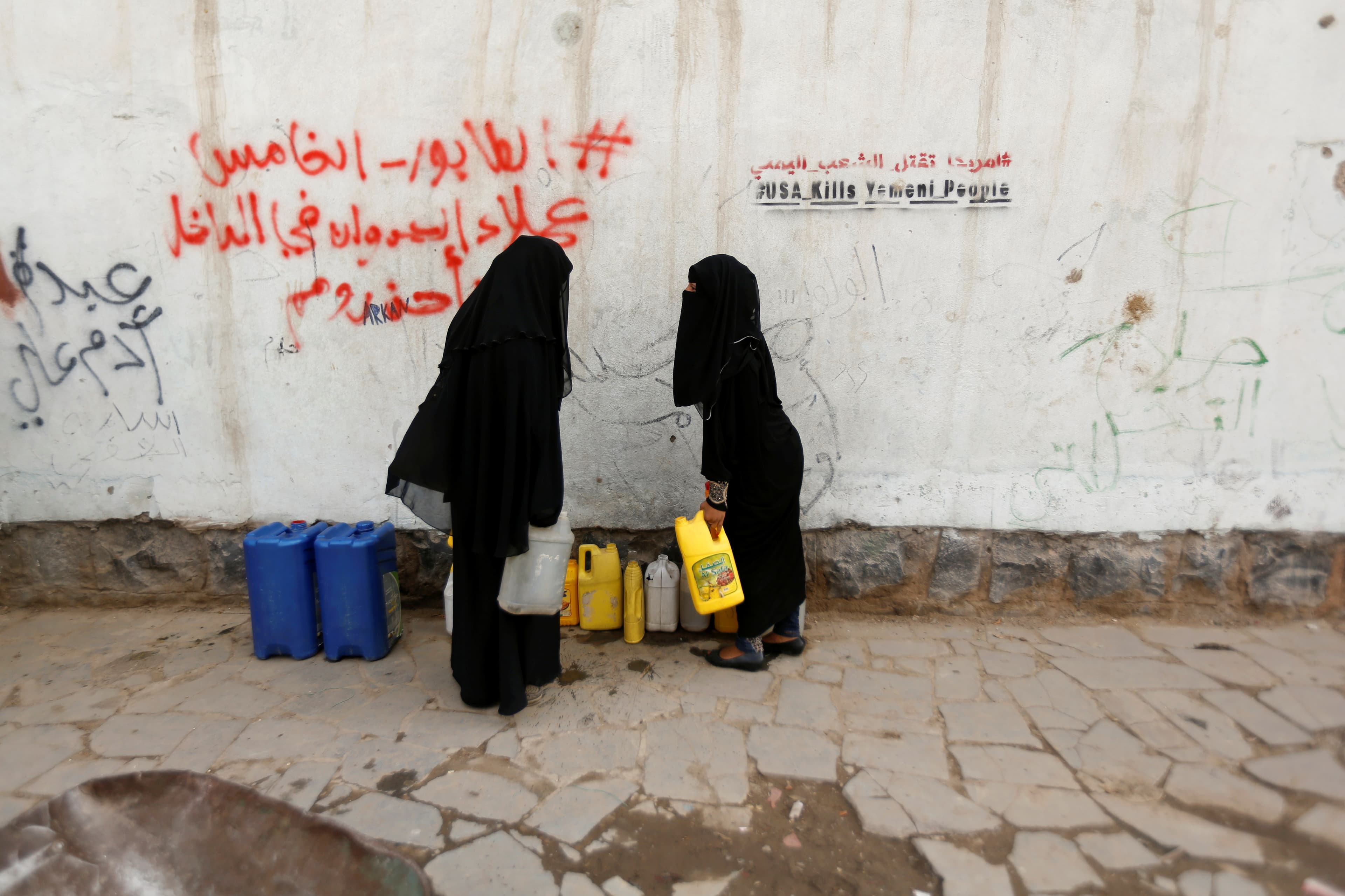 Women fully covered in black carry jerrycans after they filled them up with drinking water from a charity tap, amid a cholera outbreak, in Sanaa. Graffiti on wall behind them reads "USA kills Yemeni people."