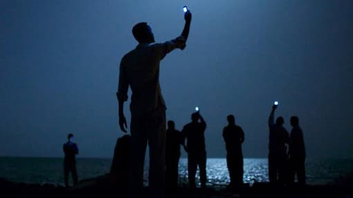 African migrants on the shore of Djibouti city at night, raising their phones in an attempt to capture an inexpensive signal from neighboring Somalia—a tenuous link to relatives abroad.