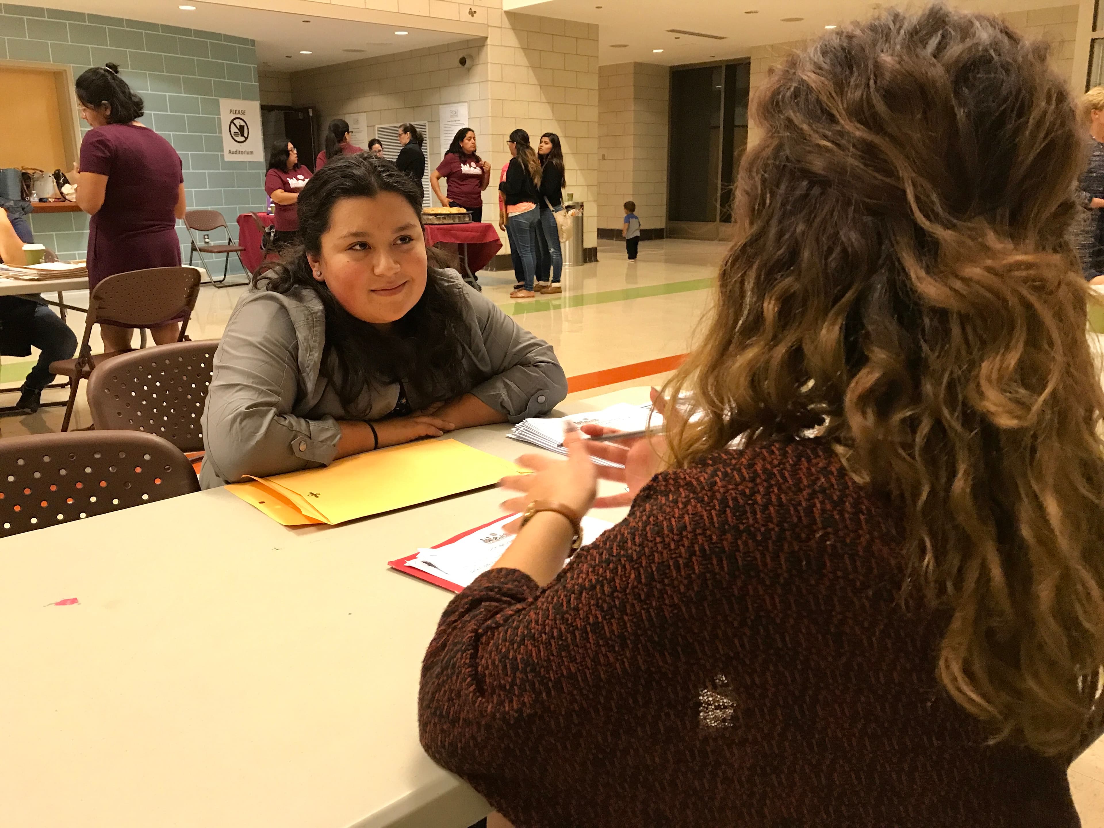 Woman sitting with papers at table in large space, another's woman's back pictured facing her