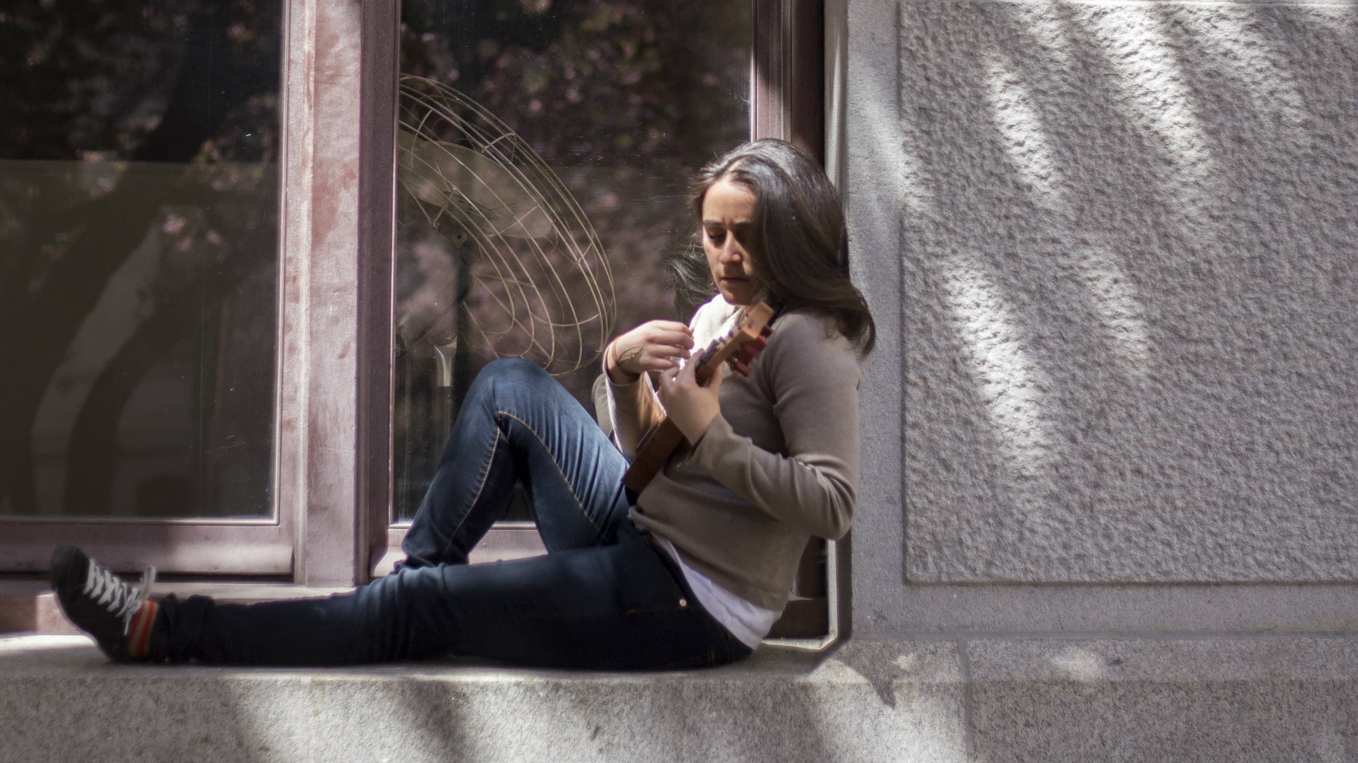 A woman sits on a window sill playing the jarana