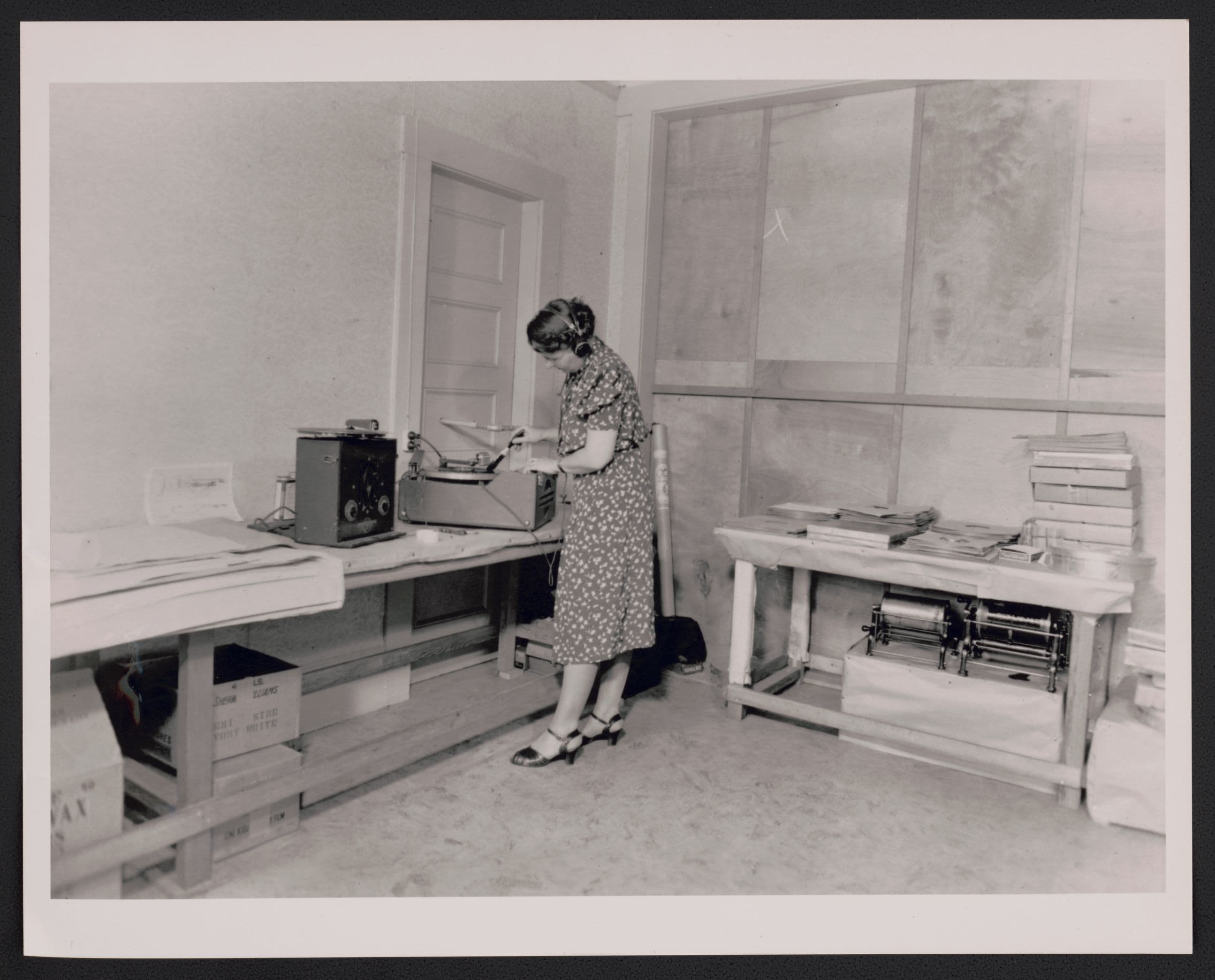 Black and white photo of woman at tabel, facing away from camera, bending over recording device