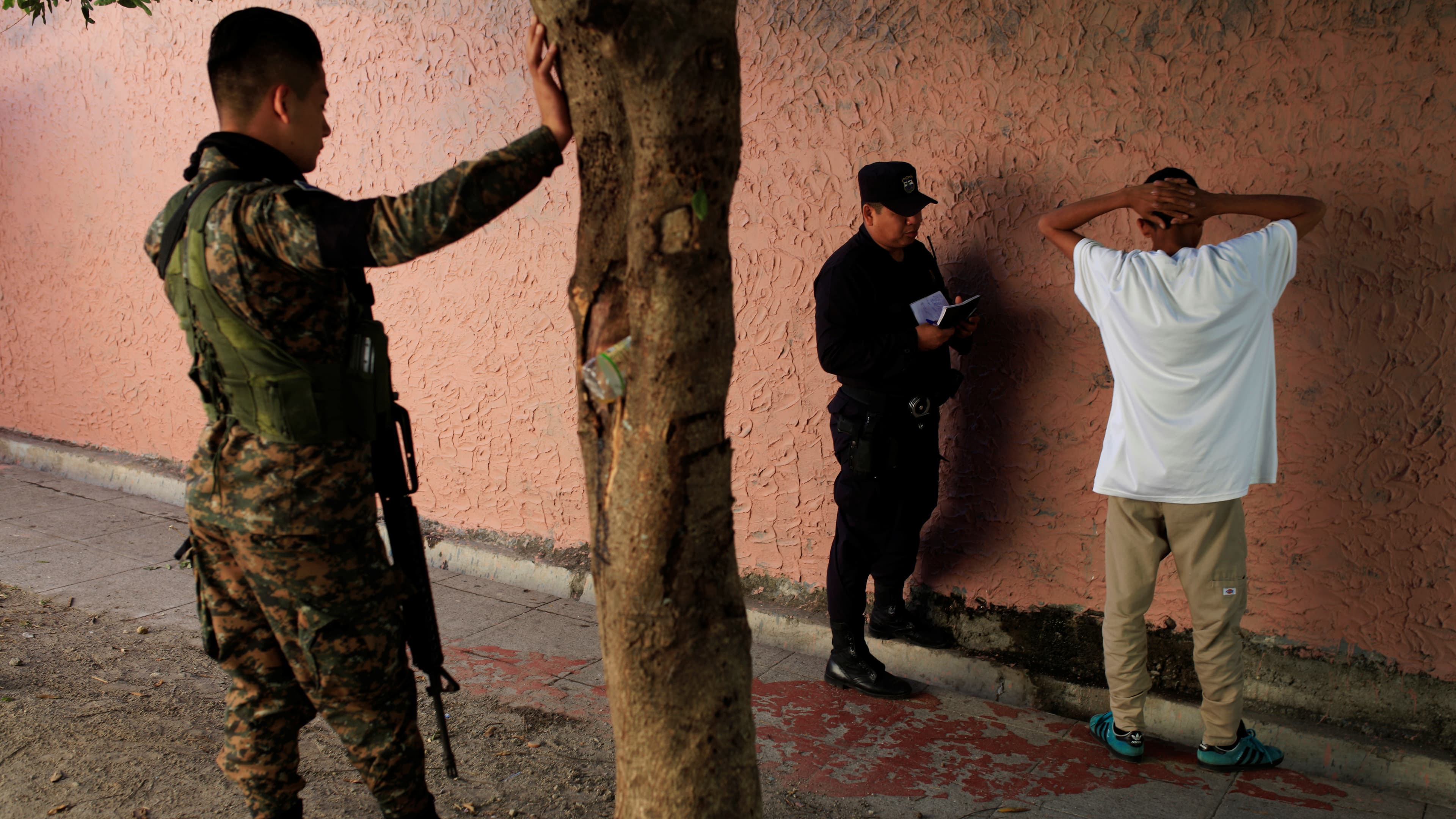 Police interrogate a suspected gang member against a wall with his hands behind his head.