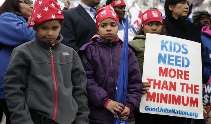 Three children stand together outside the US capitol holding a sign that reads "Kids need more than the minimum, GoodJobsNation.org."