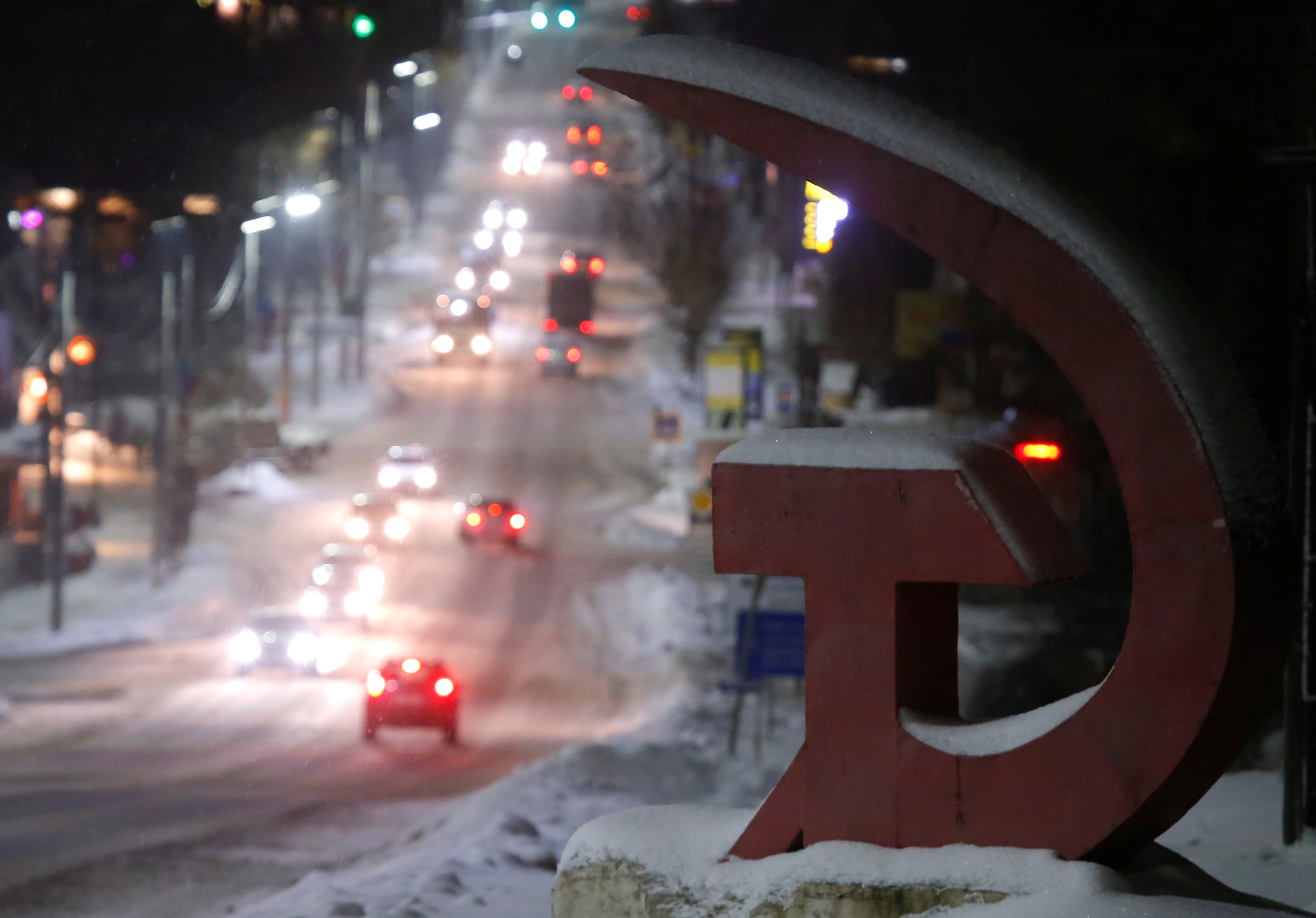 Vehicles drive past a monument covered with snow and forming a sickle and a hammer