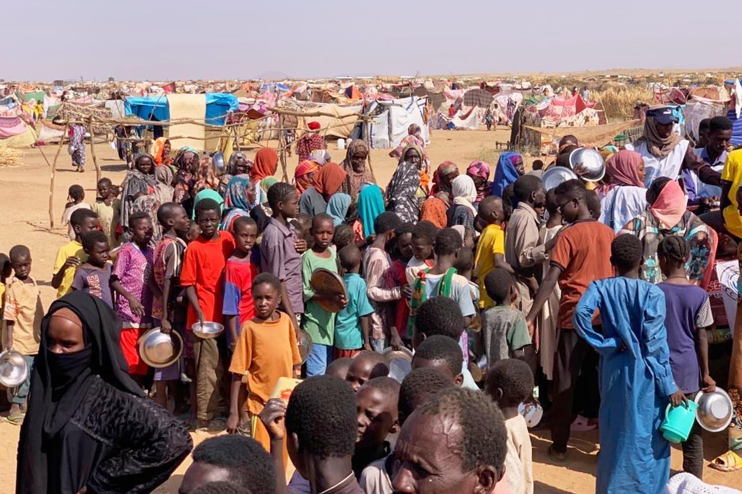 A large group of people, including many children holding metal bowls, gather in a refugee camp with makeshift tents in the background.