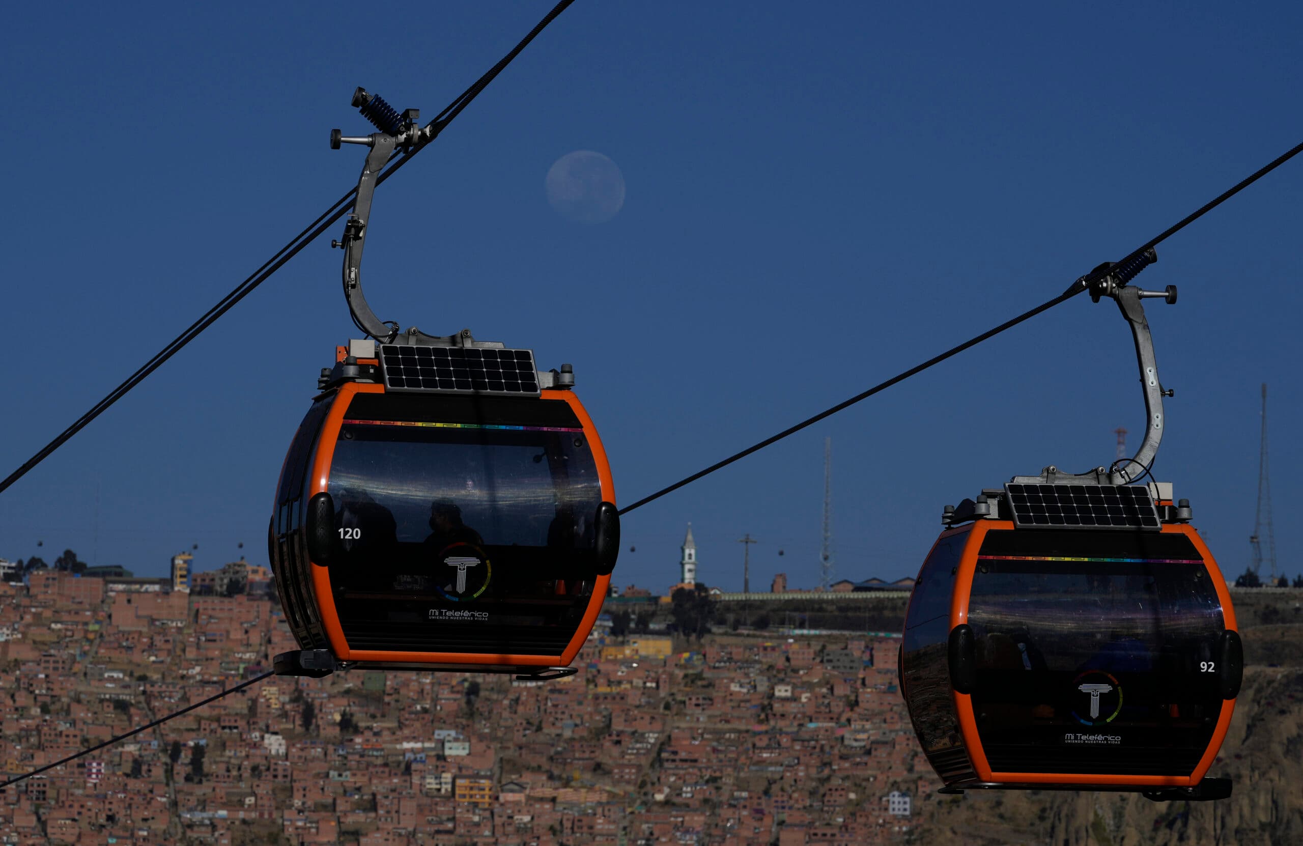 Two orange cable cars from "Mi Teleférico" gliding across a cable against a clear blue sky with a moon visible, set above a densely built hillside cityscape.