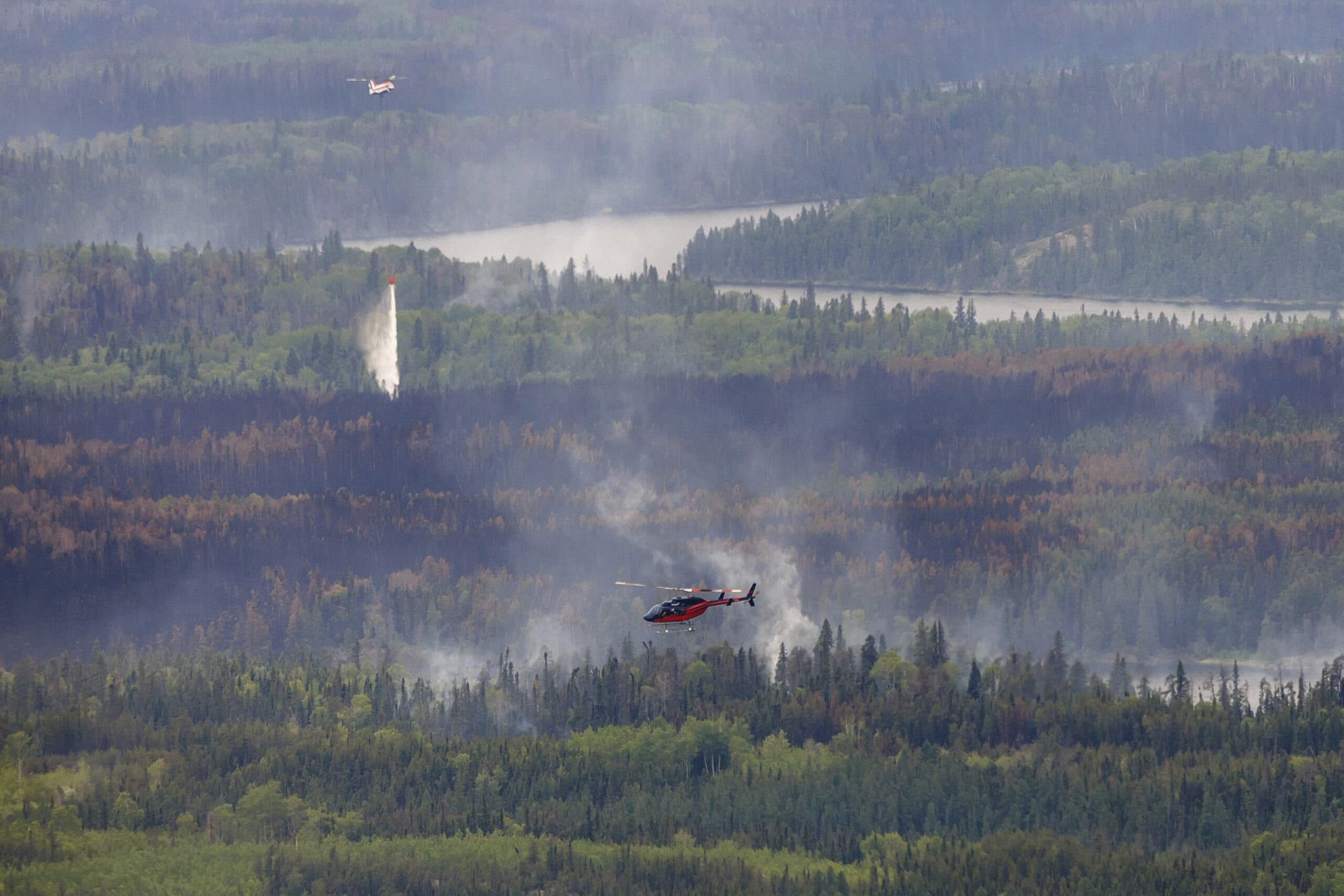 A helicopter and an aircraft engage in firefighting efforts above a forested area with visible smoke and patches of burnt trees.
