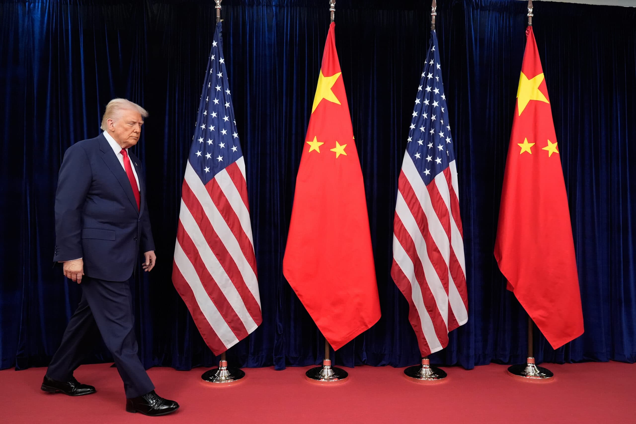 A man in a blue suit and red tie walks past a row of alternating United States and Chinese flags against a dark blue curtain backdrop.
