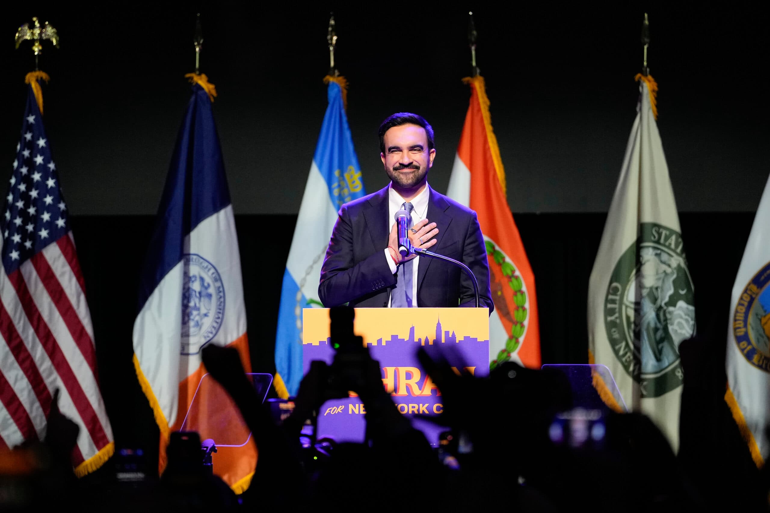 A person in a suit stands at a podium with a hand on their chest, smiling, flanked by five flags, including the American flag, during a public speaking event.