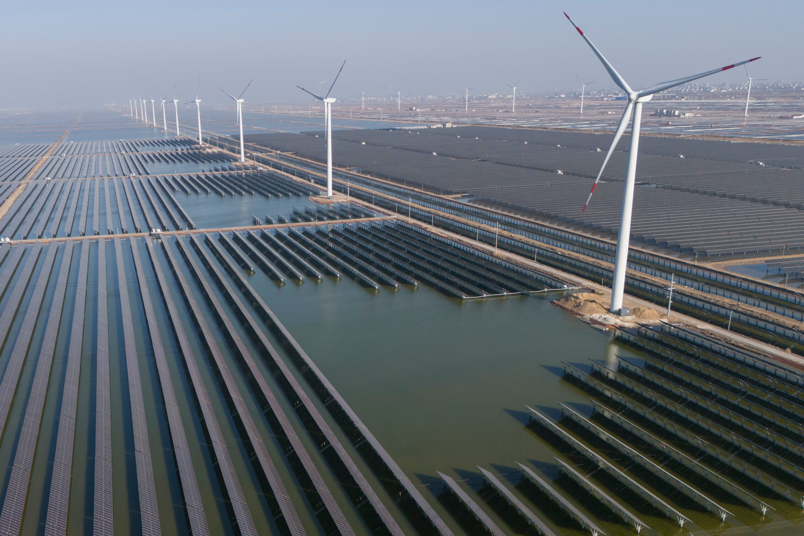 Aerial view of a large solar and wind power farm with rows of solar panels lining the ground and a series of wind turbines towering overhead, set in a landscape transitioning from land to water.