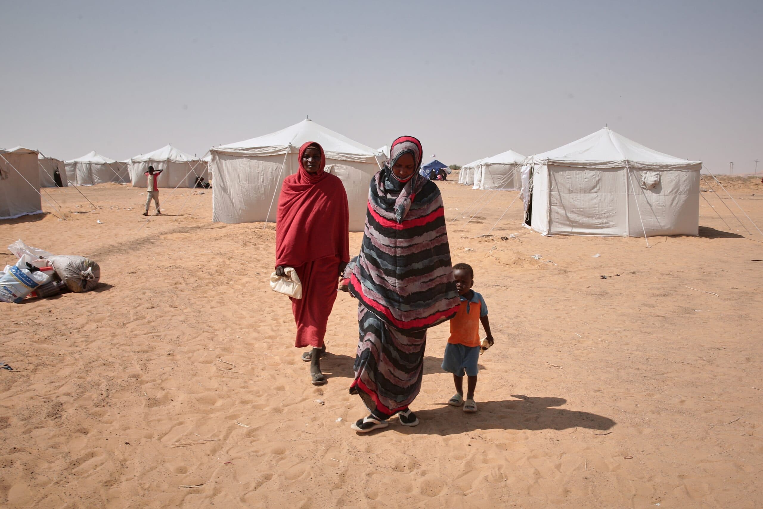 A woman and a child walk through a sandy area with several white tents in the background, while another person appears behind them in similar attire.