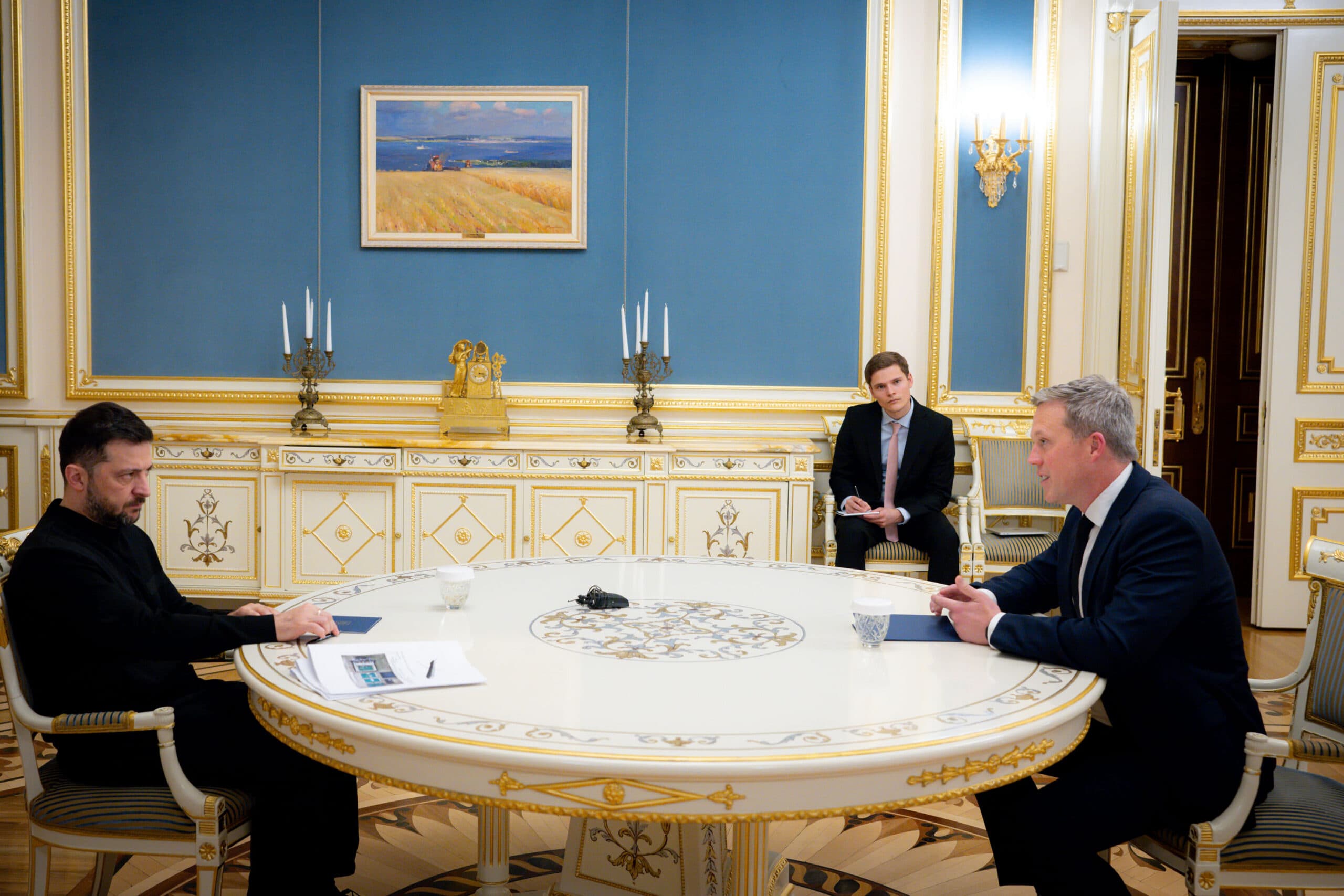 Three men sitting in a luxuriously decorated room, with ornate blue walls and golden details, engaged in a discussion around a round table.