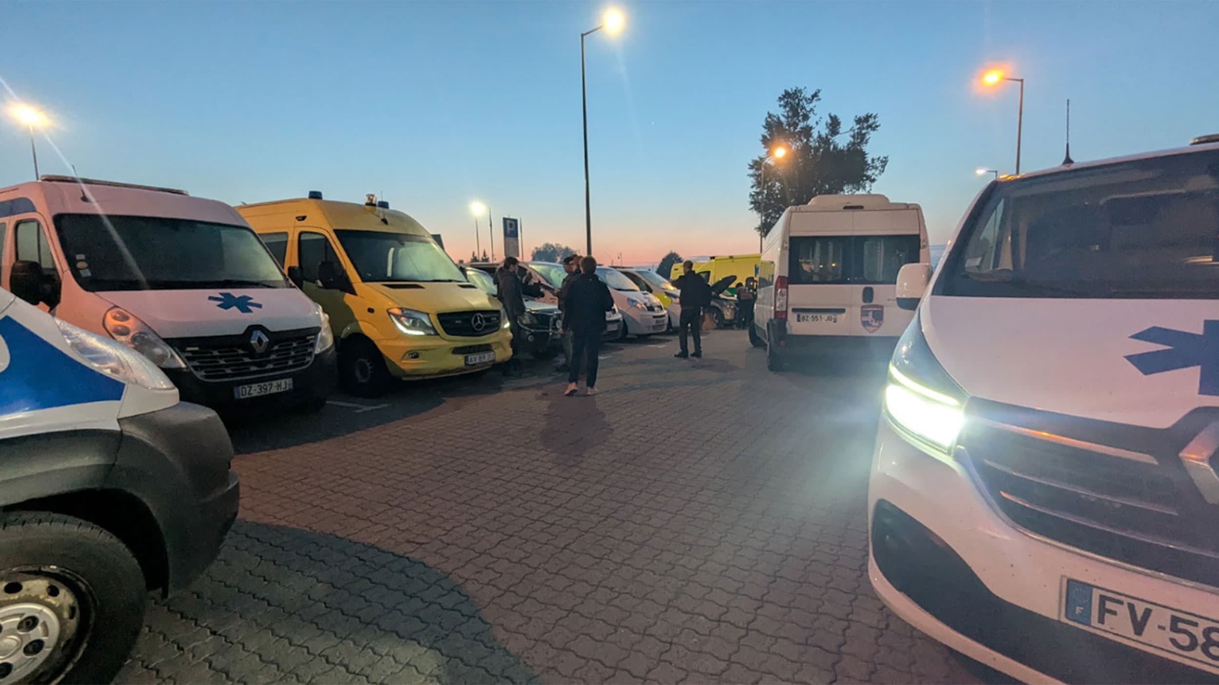 A group of ambulances parked in a lot during twilight, with a few people standing nearby under streetlights.