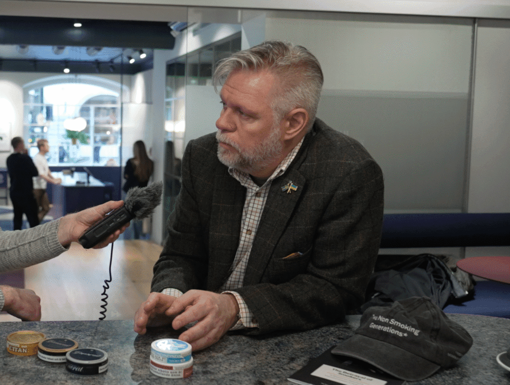 A man with gray hair and a beard, wearing a dark blazer and pin, sits at a table with several tins and a cap labeled 'Non Smoking Generations.' A person's hand is holding a microphone towards him, suggesting an interview setting, with a modern office environment in the background.