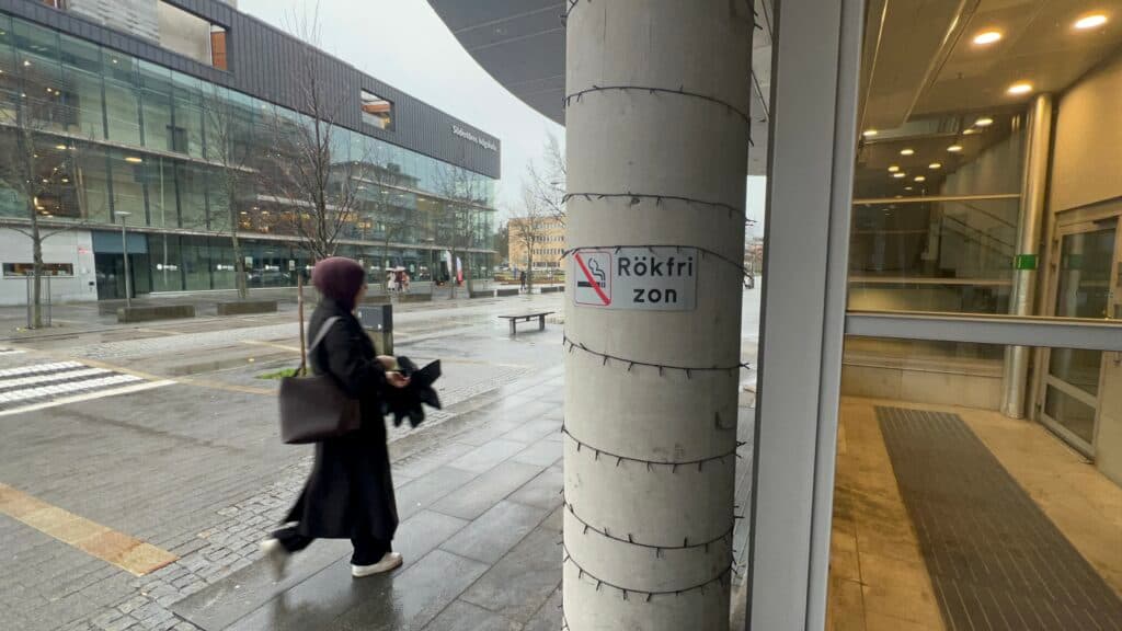 A person wearing a hijab walks past a column on a rainy day; the column is wrapped with string lights and has a "Rökfri zon" (smoke-free zone) sign.