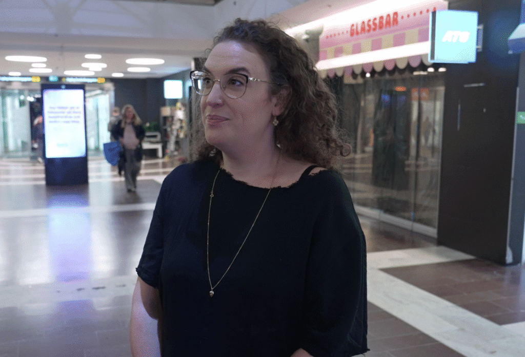A person with glasses and curly hair standing in a mall, wearing a black shirt and a long necklace. The background features a store named 'Glassbar' and a few other people walking by.